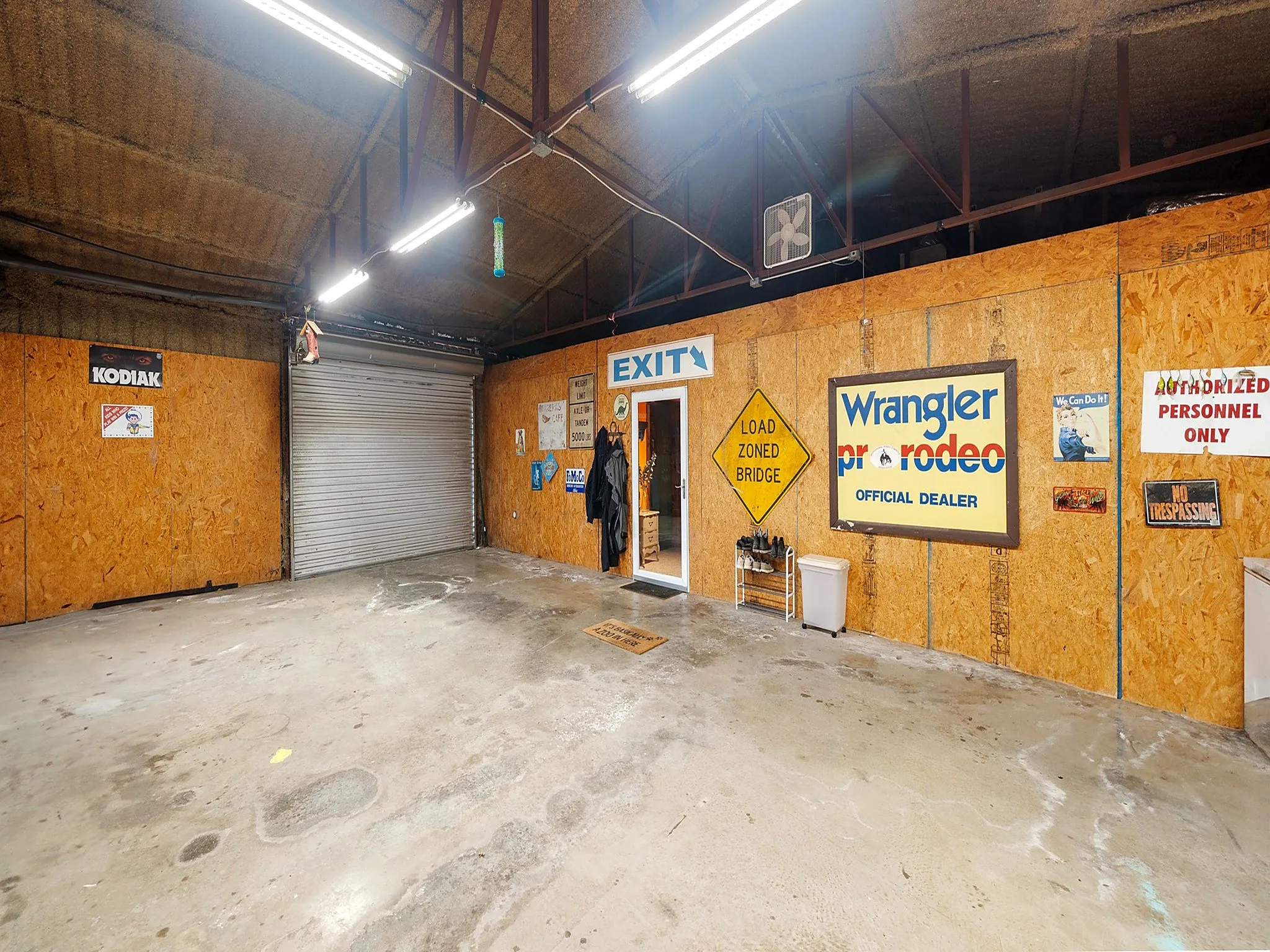 Spacious utility garage featuring exposed steel truss ceiling, concrete flooring, roll-up overhead door, and oriented strand board wall paneling