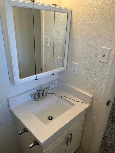 Vanity featuring a white cabinet base, quartz countertop with integrated backsplash, rectangular drop-in sink, dual-handle metal faucet, and wall-mounted tri-fold mirrored medicine cabinet