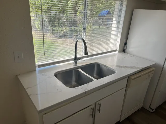 Dual-basin stainless steel sink featuring a high-arc pull-down faucet, white stone-patterned countertop, and white under-counter cabinetry with metallic hardware