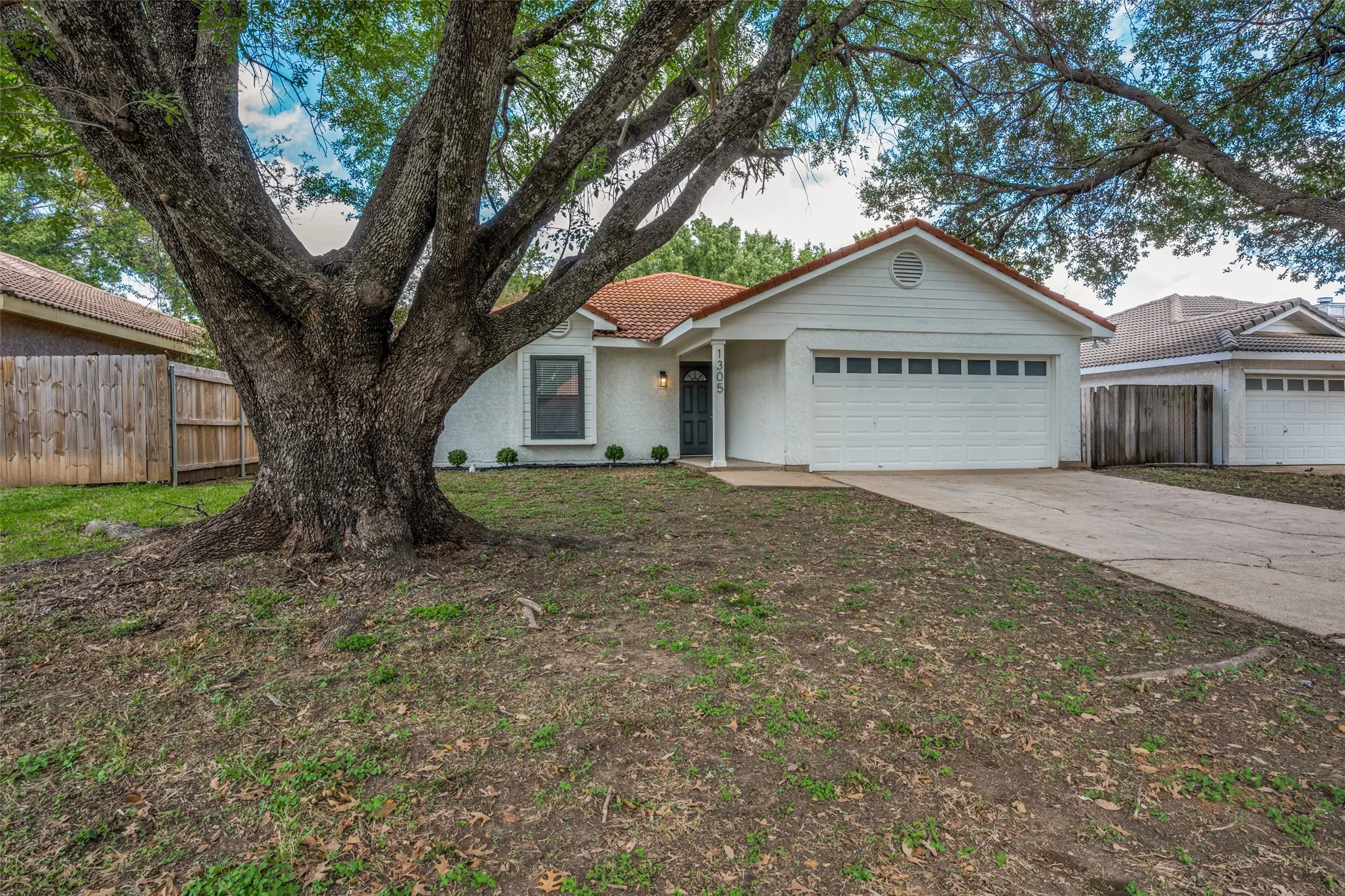 Single-story residence featuring a tile roof, stucco exterior, mature shade tree, two-car garage, and concrete driveway