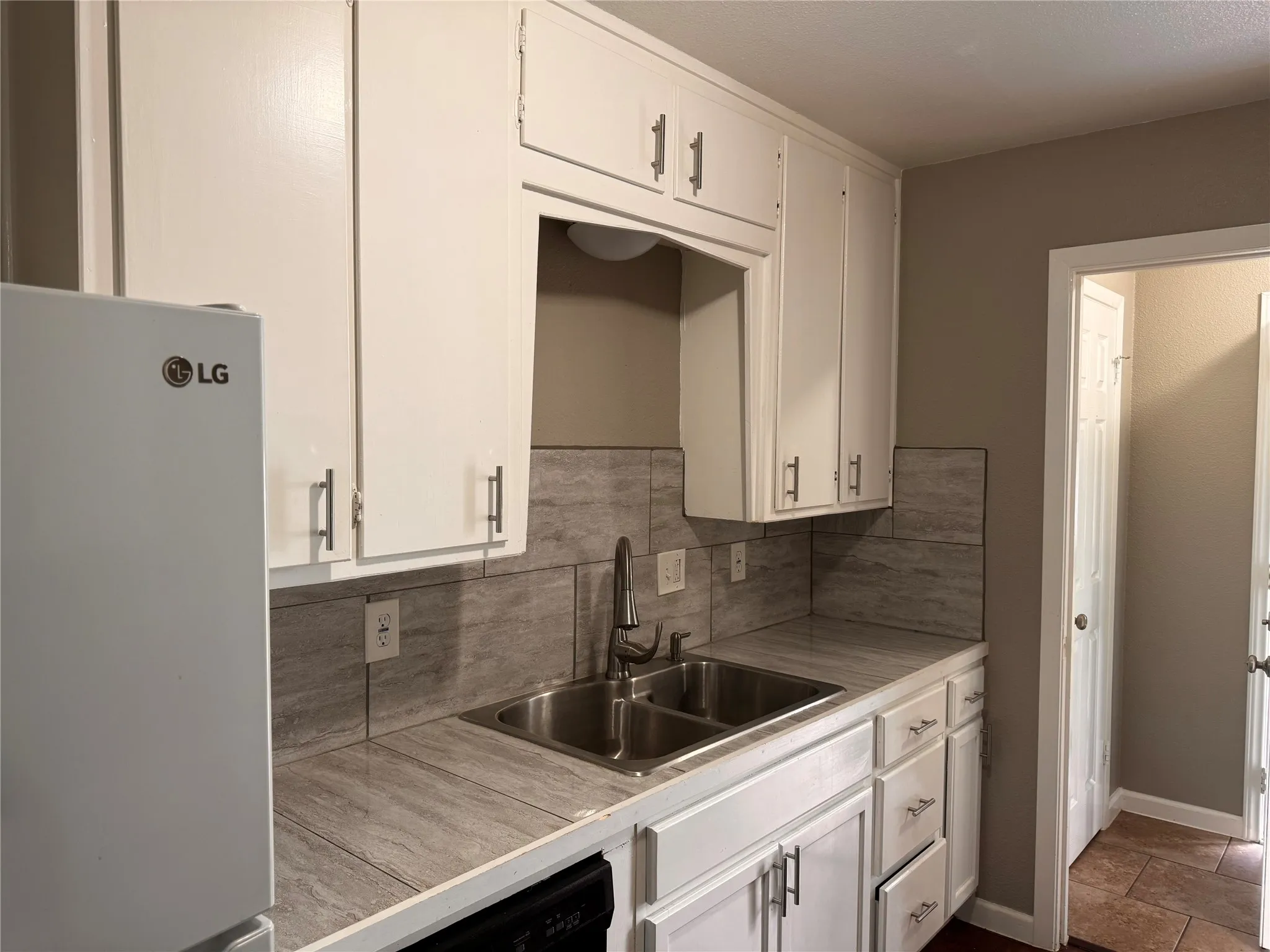 Kitchen featuring white cabinetry with metallic pulls, stone-texture tile countertops, coordinating backsplash, dual-basin stainless steel sink with a high-arch faucet, and white appliances