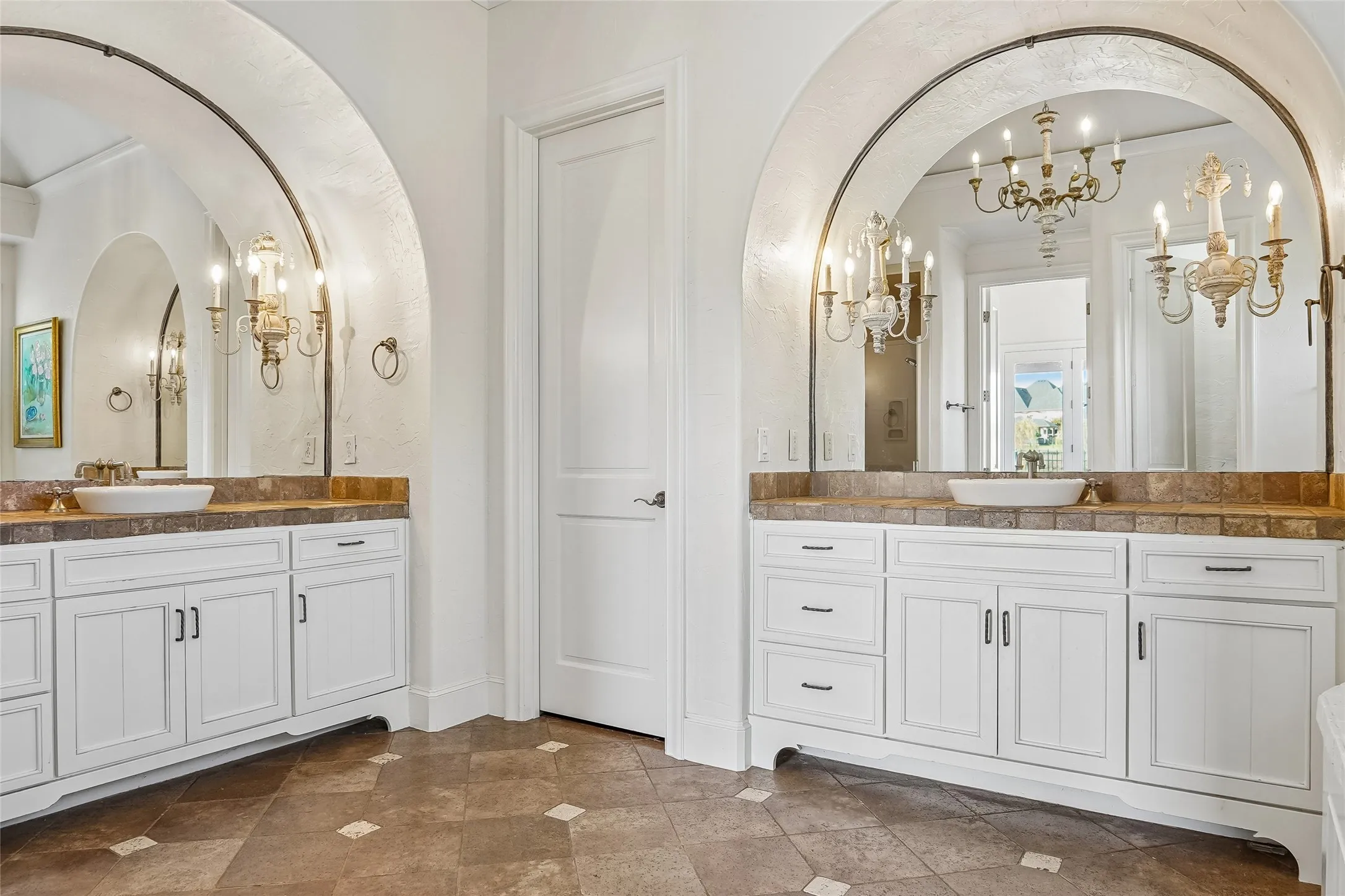 Dual white vanities featuring stone-tile countertops, integrated vessel sinks, arched mirrors, and wall-mounted multi-arm chandeliers, finished with stone-tile flooring