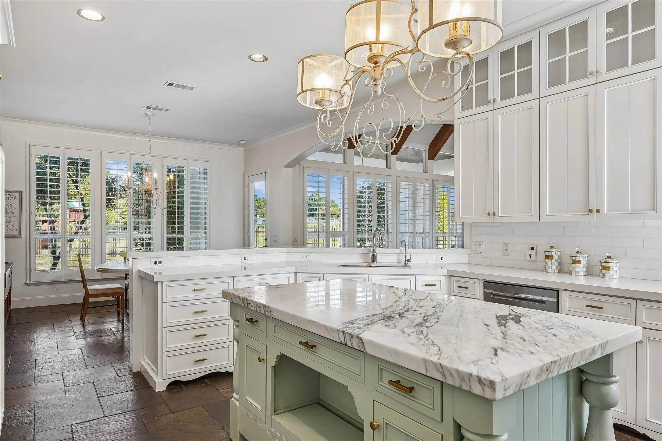 Kitchen featuring stone-topped island with base cabinetry, ceiling-mounted decorative chandelier, white shaker-style cabinetry with glass-front uppers, subway tile backsplash, and stone tile flooring