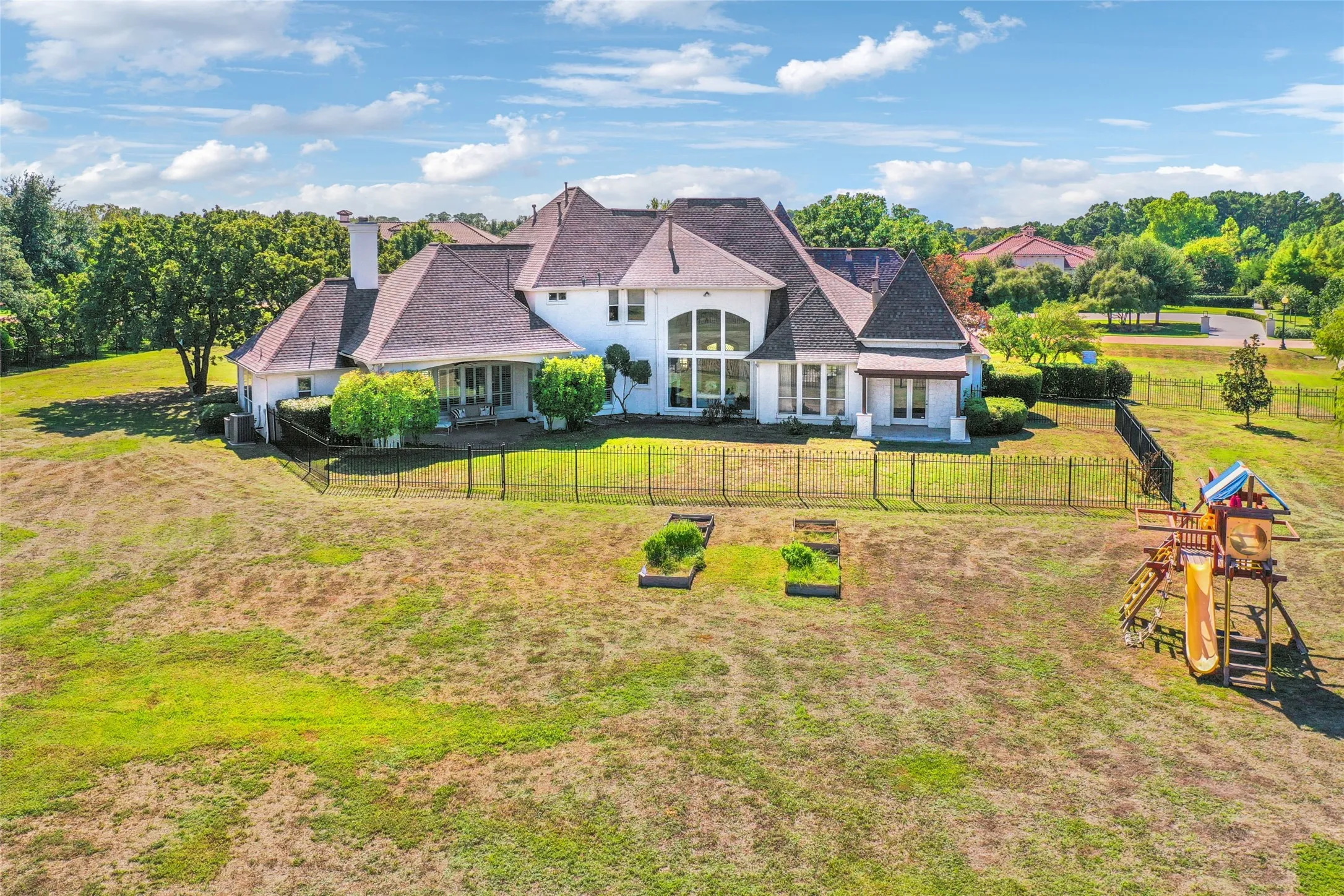 Expansive backyard featuring a wrought-iron perimeter fence, raised garden beds, and multi-gabled architectural roofline