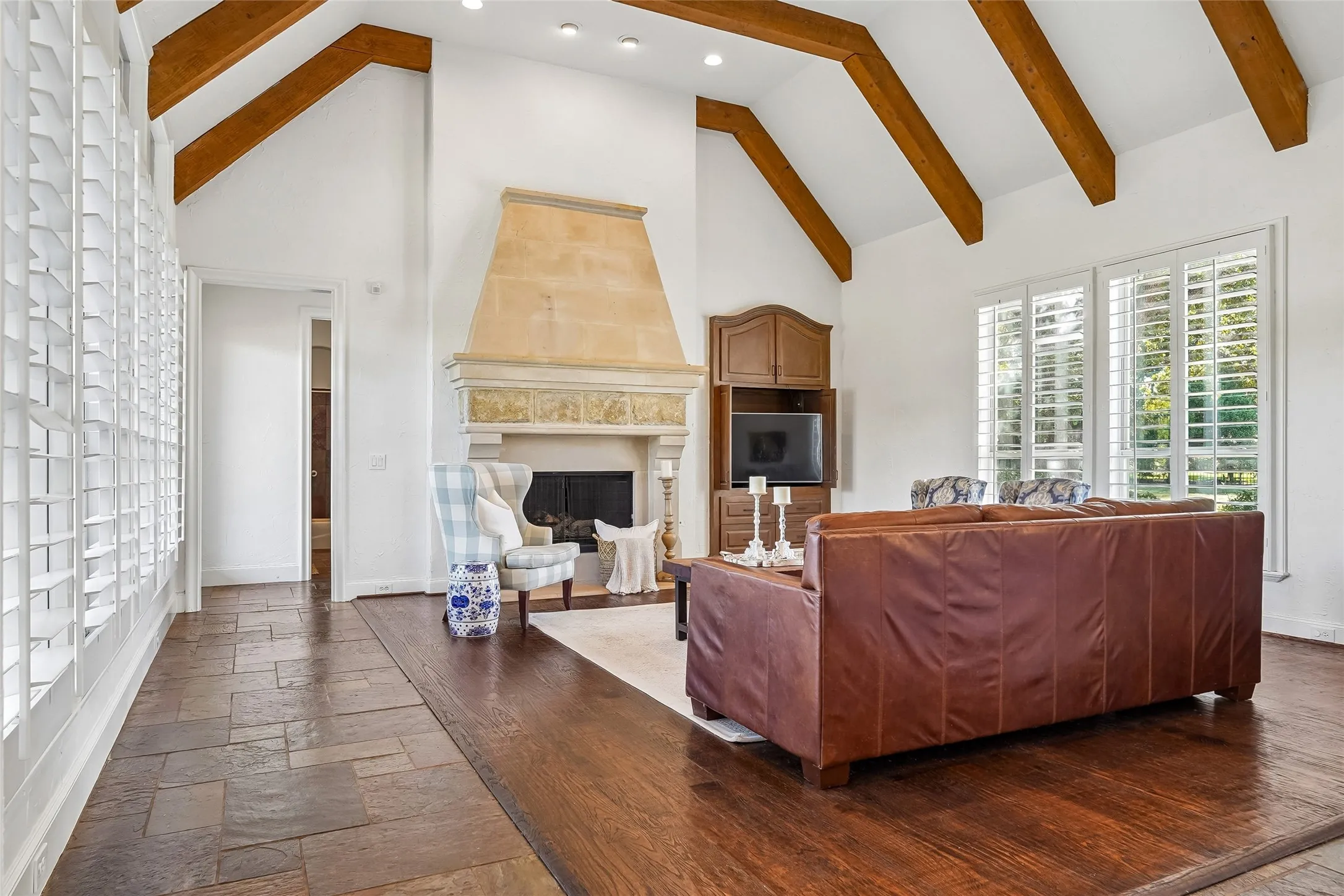 Vaulted ceiling featuring exposed wood beams, stone fireplace with a decorative mantel, wood-finish flooring, stone tile inlay, and plantation shutters