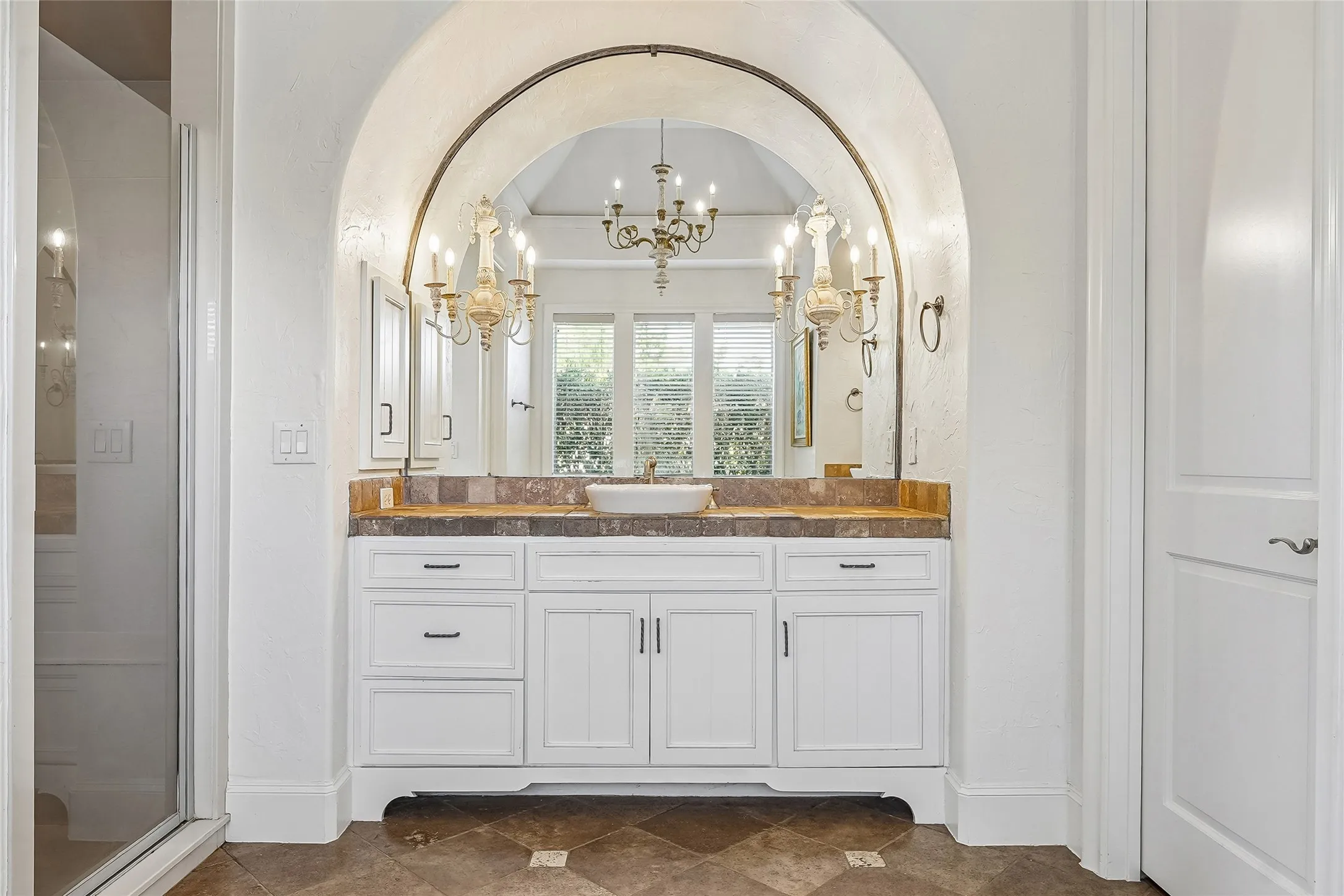 Arched vanity alcove featuring a white cabinetry base, stone tile countertop with a vessel sink, large arched wall mirror, and dual ornate light fixtures