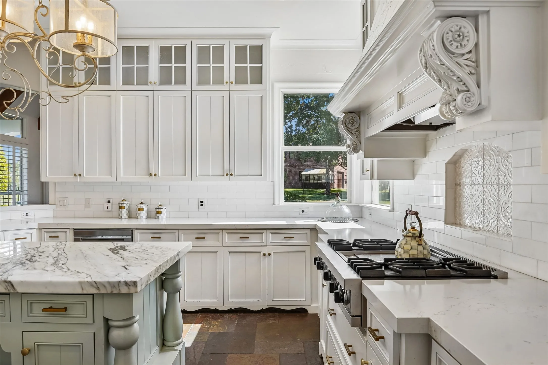 Kitchen featuring floor-to-ceiling cabinetry with glass-panel upper doors, a stone-topped island with decorative column accents, professional-grade gas range, custom range hood with corbel detailing, and a white subway tile backsplash