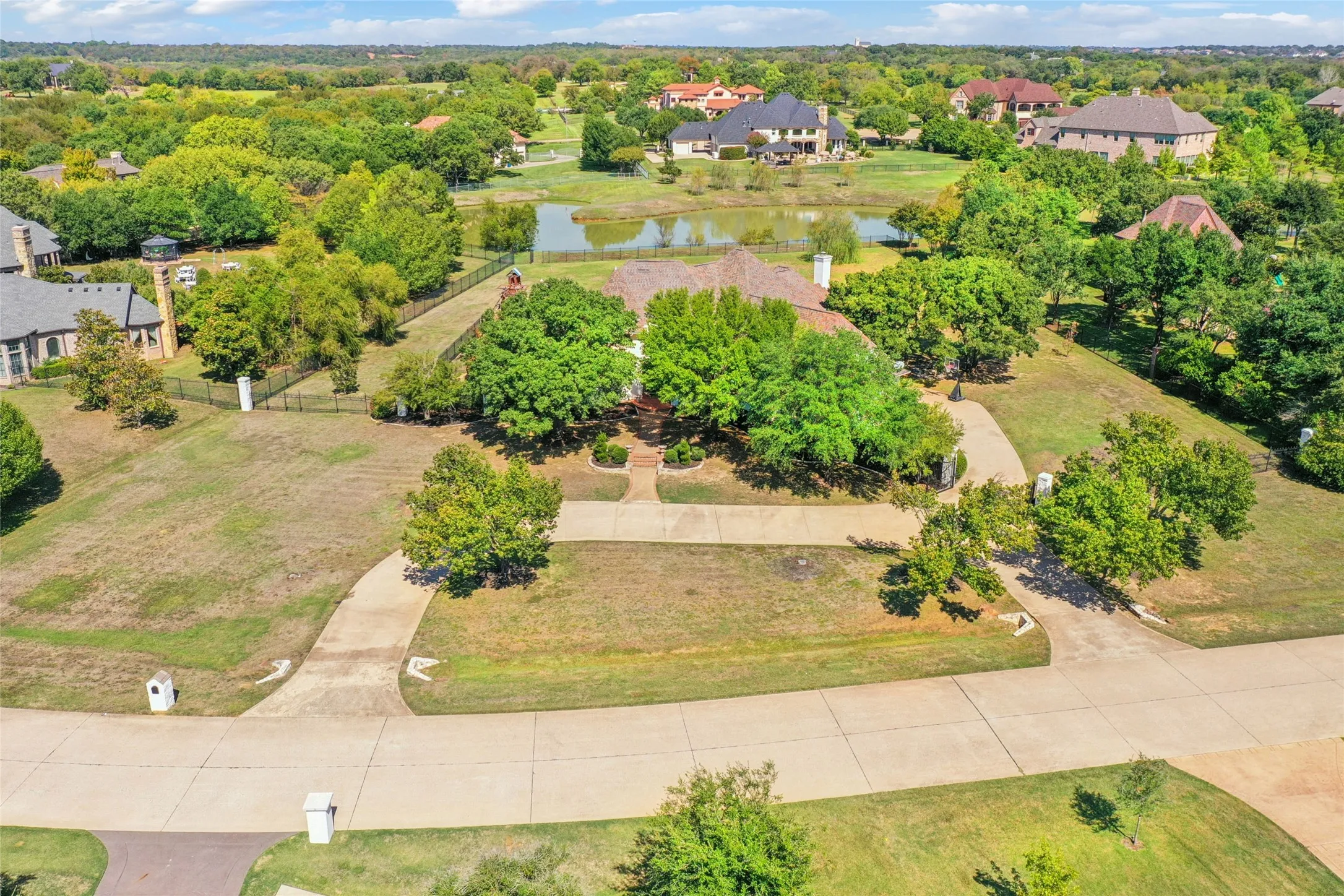 Aerial perspective of a sprawling lot featuring a multi-segment concrete driveway, mature trees, open lawn space, and fenced boundaries