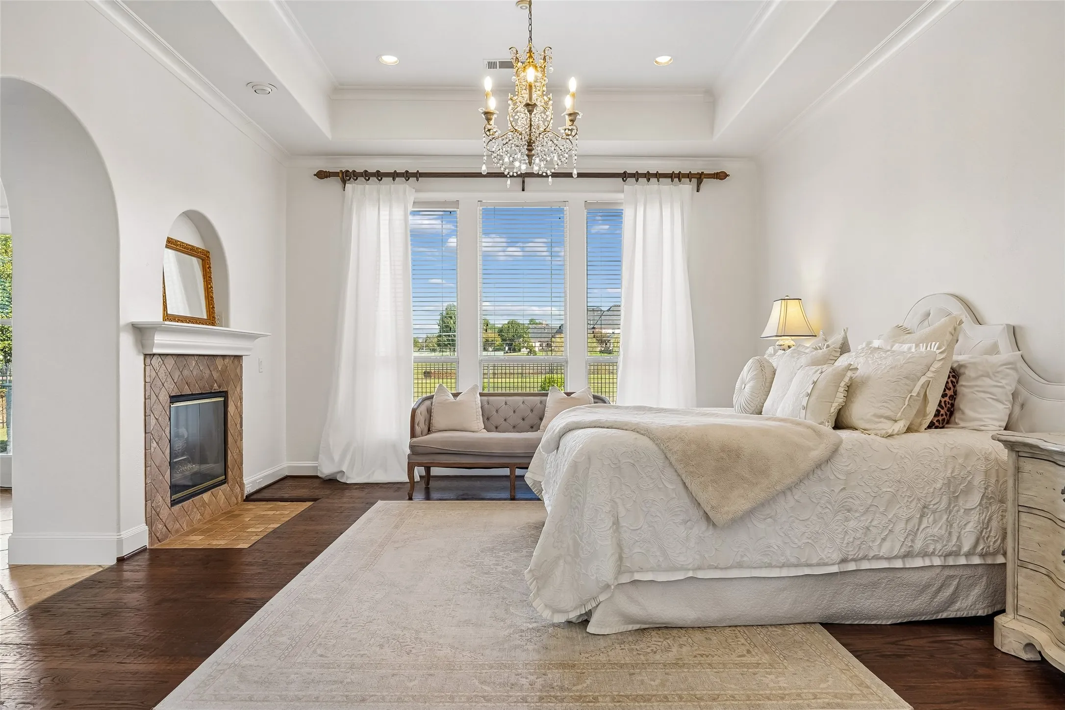 Tray ceiling with a central crystal chandelier, wood-finish flooring, and a fireplace featuring a tile surround and arched inset niche