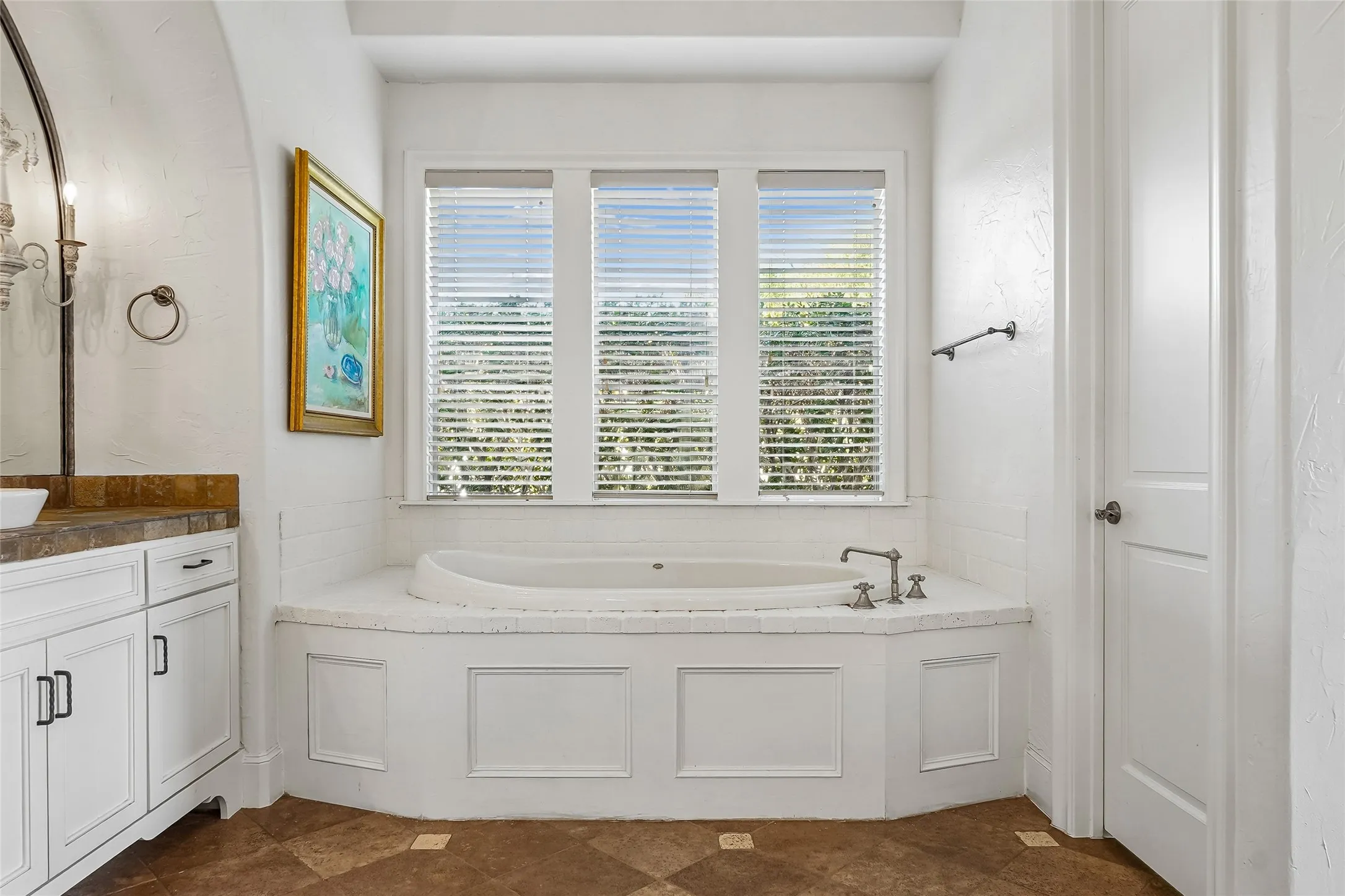 Built-in soaking tub with a custom white paneled surround and stone tile deck, complemented by a triple-window configuration, white cabinetry with stone countertop, and tile flooring