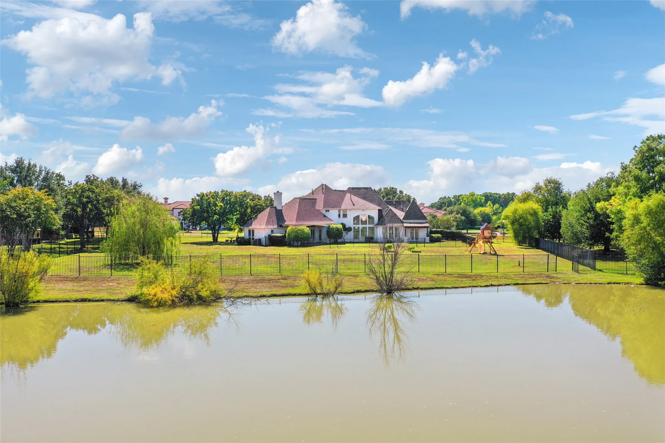 Multi-story residence featuring a complex hip roofline, exterior chimney, expansive rear lawn, perimeter metal fencing, and water frontage