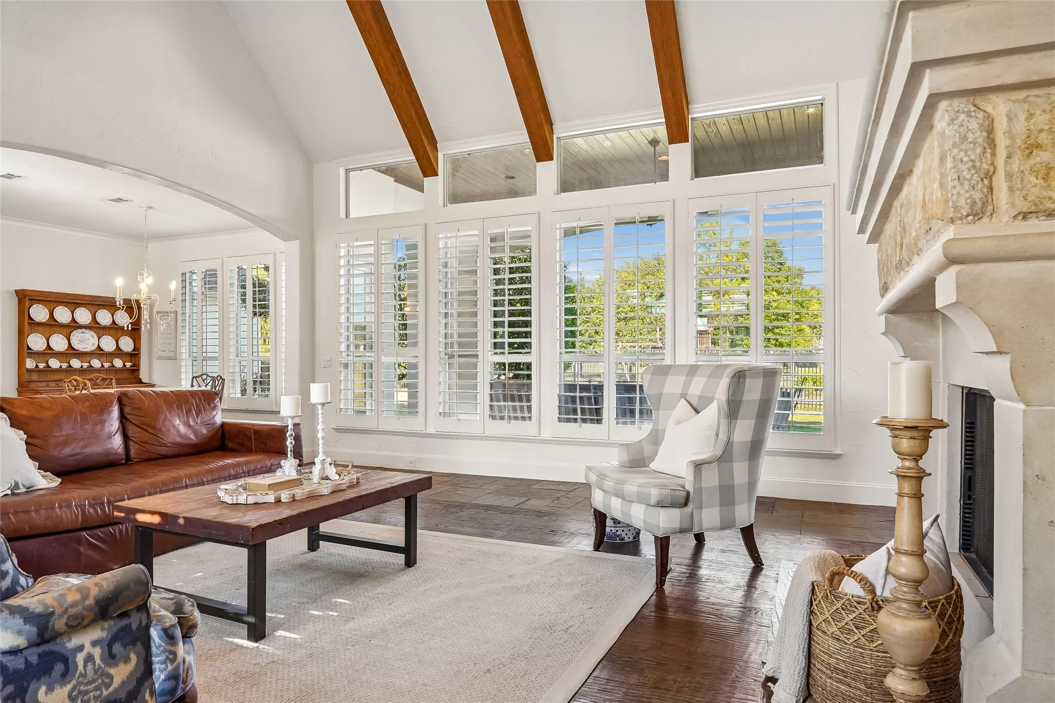 Vaulted ceiling featuring exposed wood beams, stone-surround fireplace, and floor-to-ceiling windows with plantation shutters