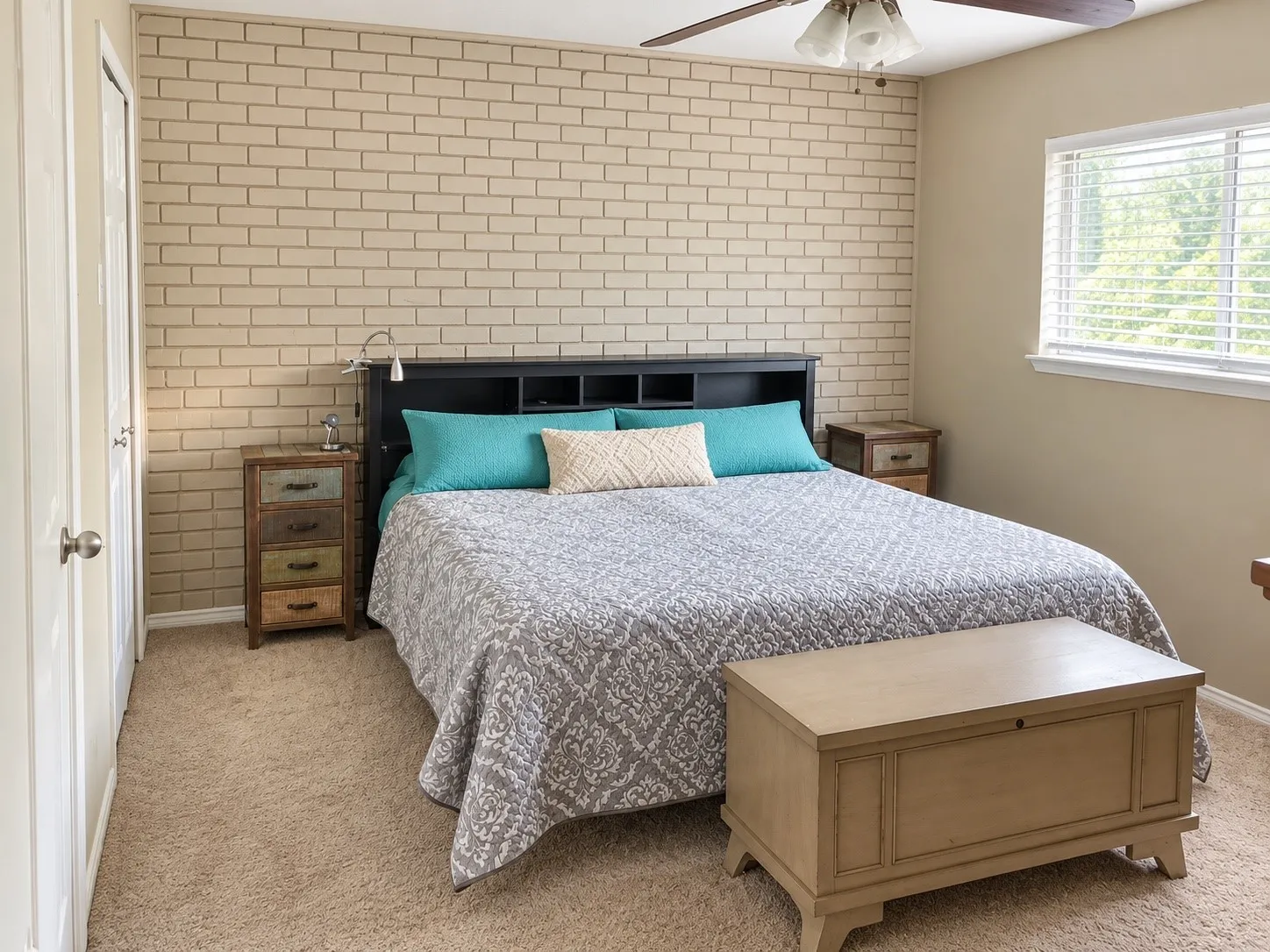 Neutral-toned brick accent wall, carpeted flooring, window with horizontal blinds, and ceiling fan with integrated lighting