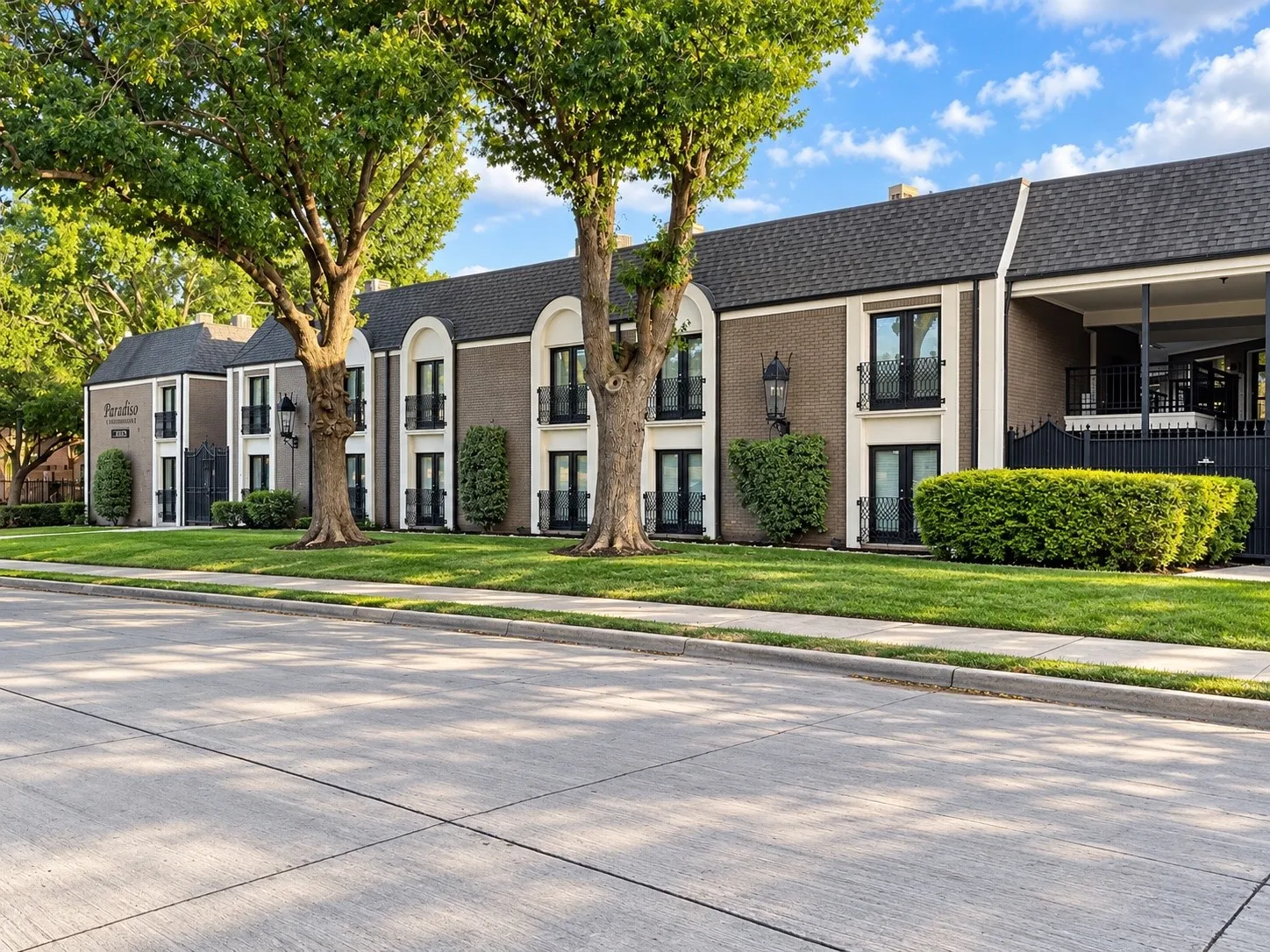 Multi-story brick exterior featuring arched architectural accents, wrought iron balcony railings, dark shingled roof, and mature landscaping
