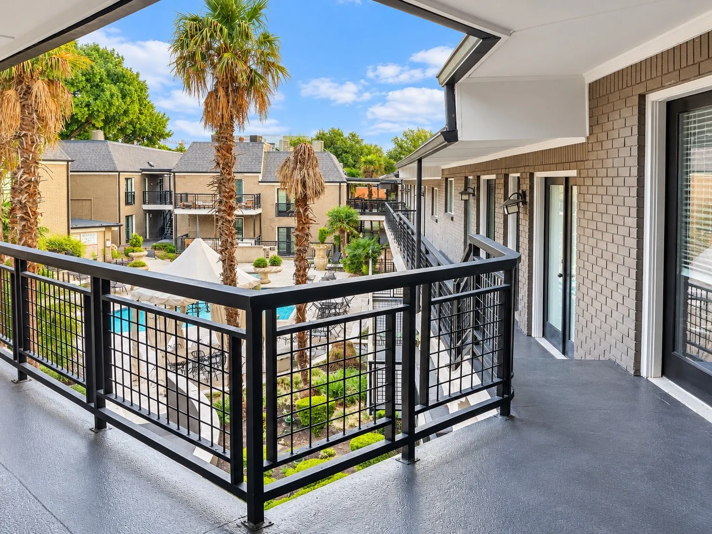 Covered balcony featuring black metal railings, textured concrete flooring, painted brick exterior, and wall-mounted exterior lighting