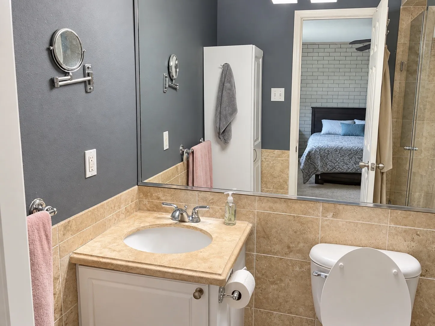 Bathroom featuring a white vanity cabinet, neutral stone-finish countertop with an integrated basin, large-format tile wainscoting, wall-to-wall vanity mirror, and built-in vertical storage cabinetry