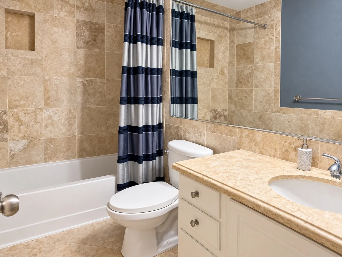 Bathroom featuring full-height wall tiling, built-in recessed shower shelving, a vanity with a stone-finish countertop, an oval under-mount sink, and a full-width wall mirror