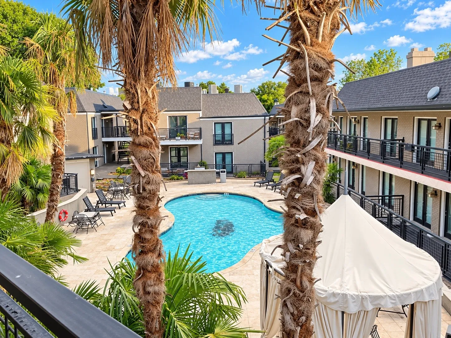 Swimming pool with surrounding stone-finished patio, mature palm trees, and multi-story buildings featuring exterior metal railings