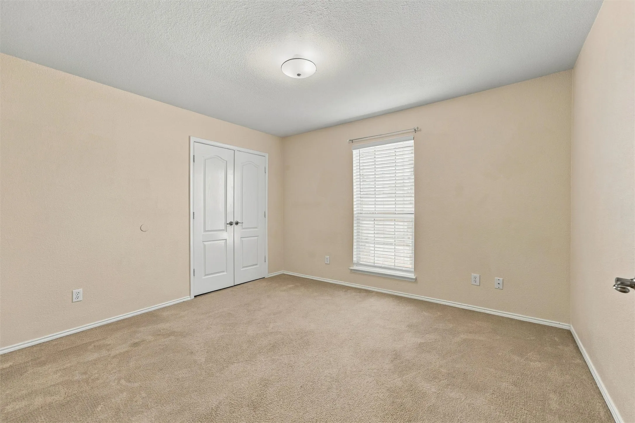 Interior room featuring neutral-toned wall paint, plush carpeting, double-panel closet doors, a single window with integrated horizontal blinds, and a flush-mount ceiling light fixture