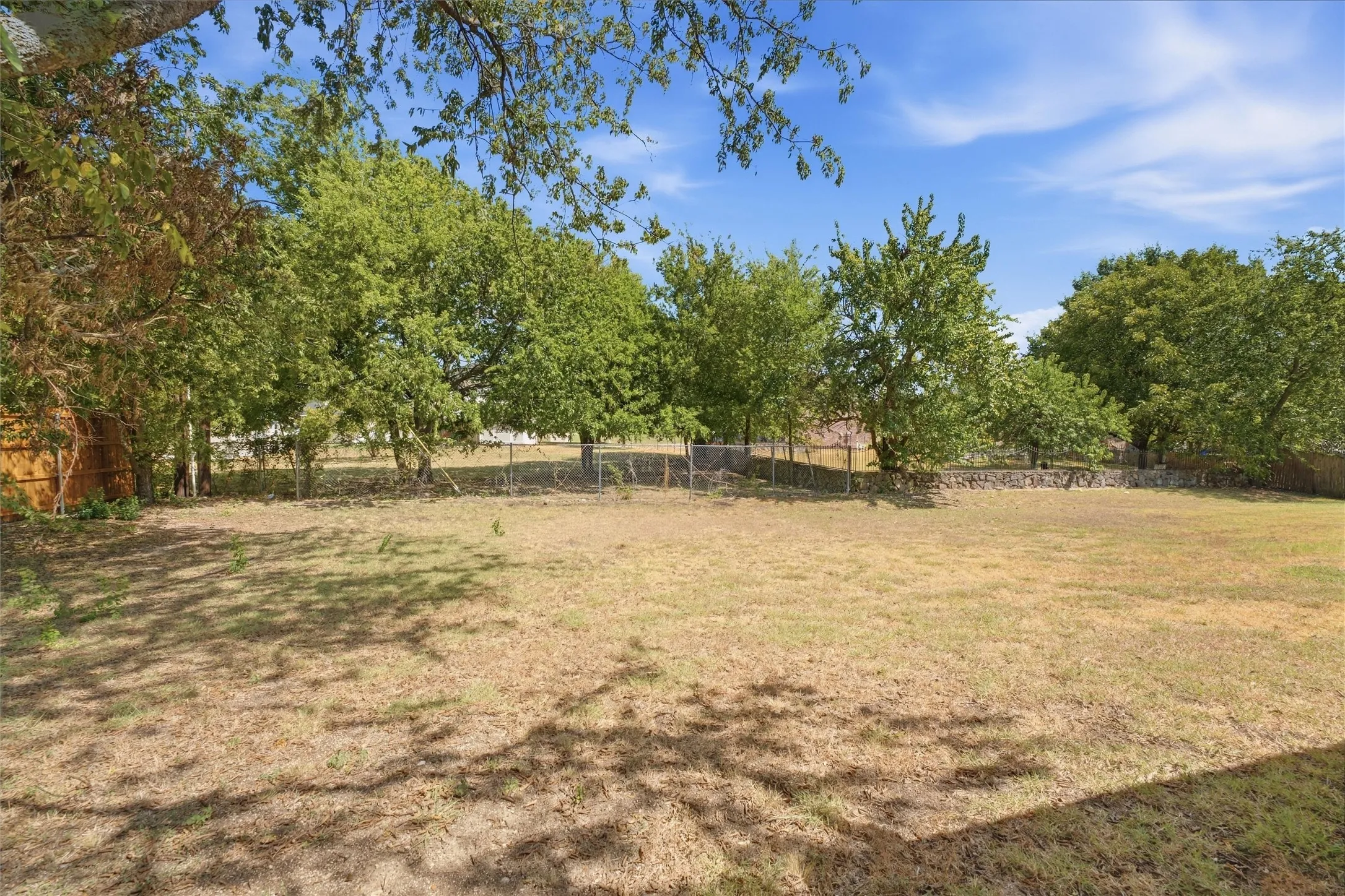 Expansive backyard featuring a fenced perimeter, mature shade trees, and stone masonry wall accents