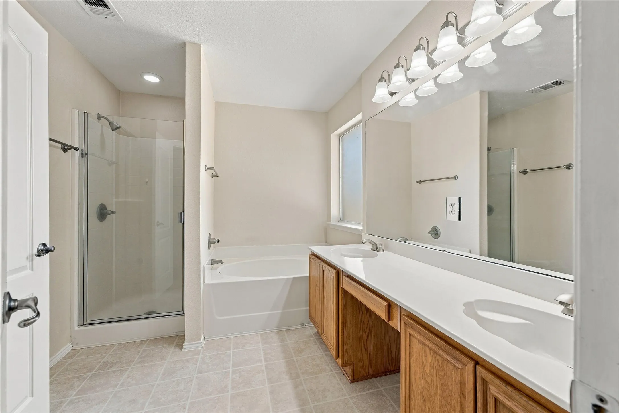 Bathroom featuring a dual-sink vanity with a solid-surface countertop, full-width vanity mirror, separate glass-enclosed shower, and a built-in soaking tub