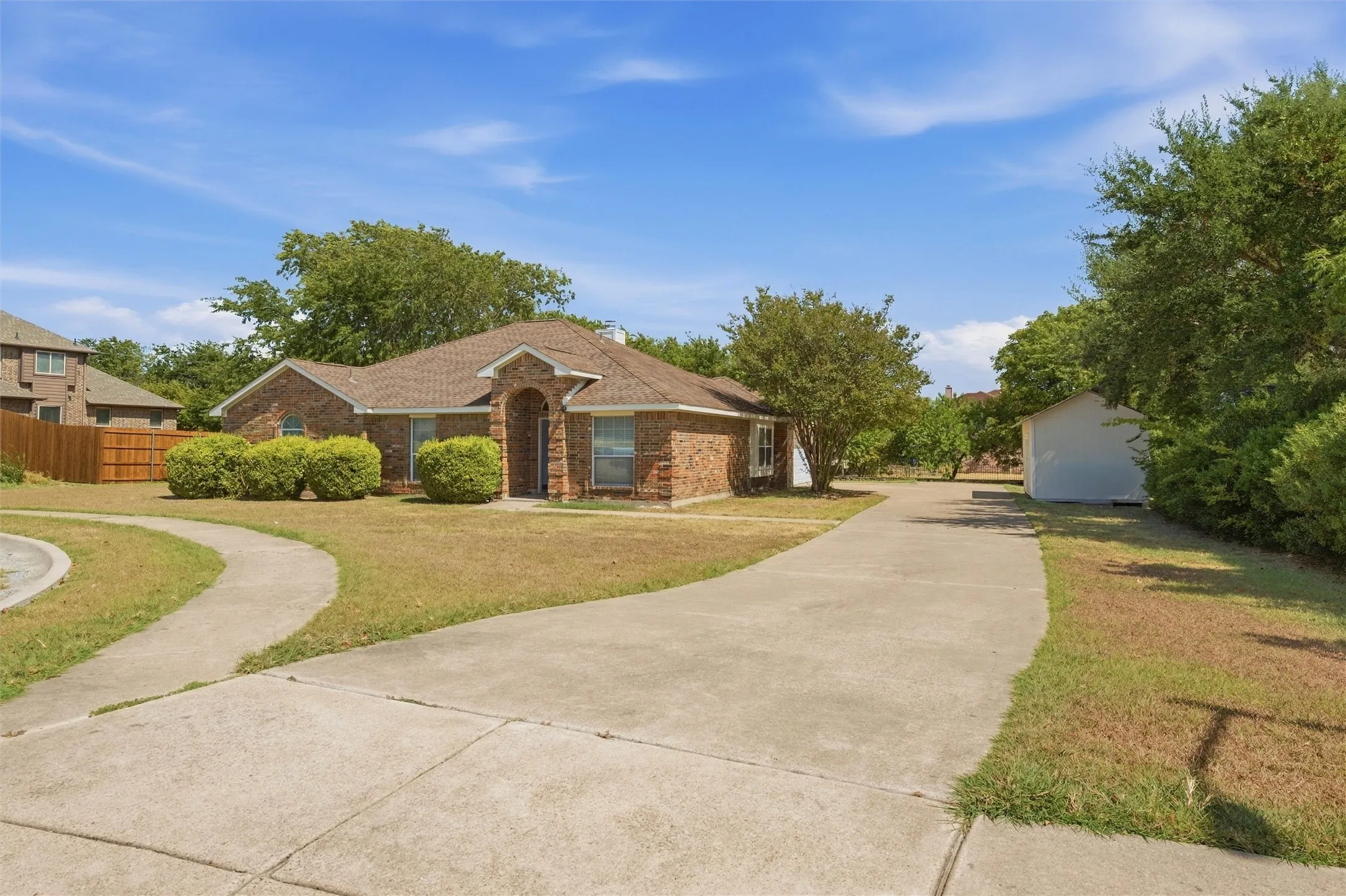 Brick exterior residence featuring an arched entryway, a sprawling concrete driveway, and a detached storage structure set on a manicured lot