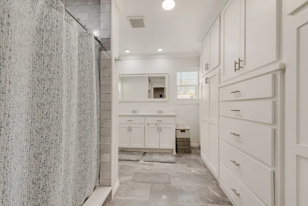 Bathroom featuring large-format tile flooring, extensive white built-in cabinetry with brushed nickel hardware, a dual-sink vanity, and a tub enclosure with subway-tiled walls
