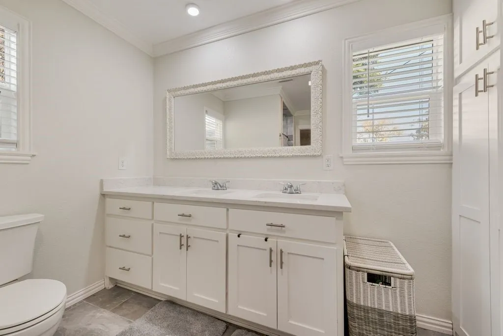 Dual-sink vanity with a stone-surface countertop, white shaker-style cabinetry, crown molding, framed wall mirror, and integrated floor-to-ceiling storage cabinetry