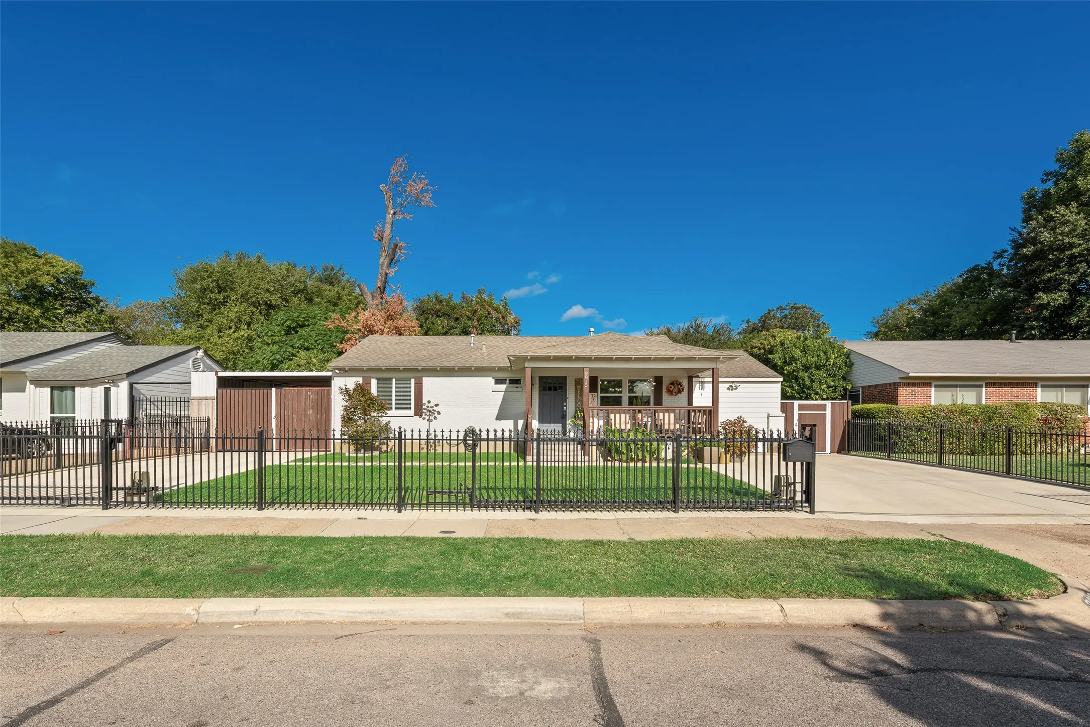 Single-story residence featuring a light-colored exterior, covered front porch with a wood-finished railing, metal perimeter fencing, and an attached carport