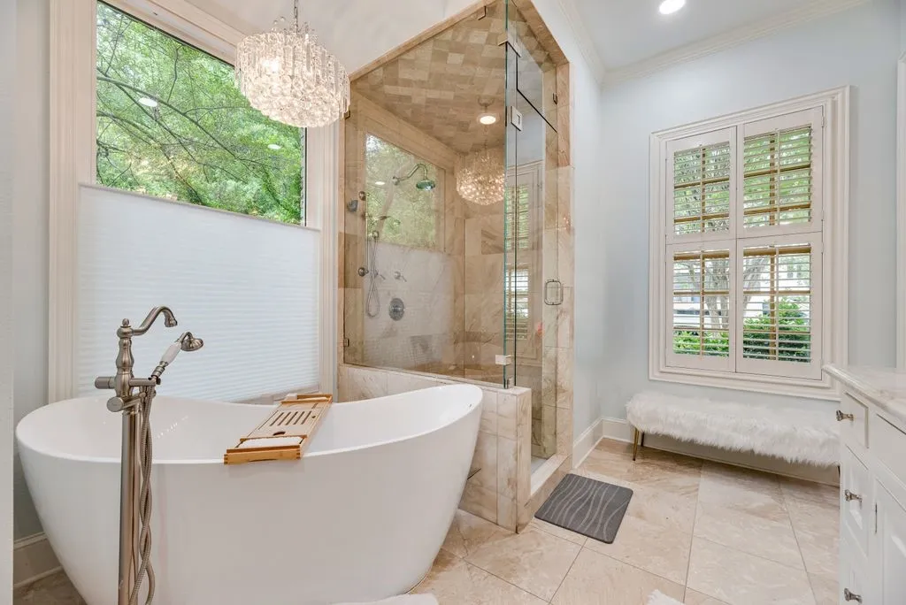 Freestanding soaking tub with floor-mounted brushed nickel faucet, glass-enclosed shower with stone tile surround, multi-pane windows with integrated shutters, and crystal chandelier fixture