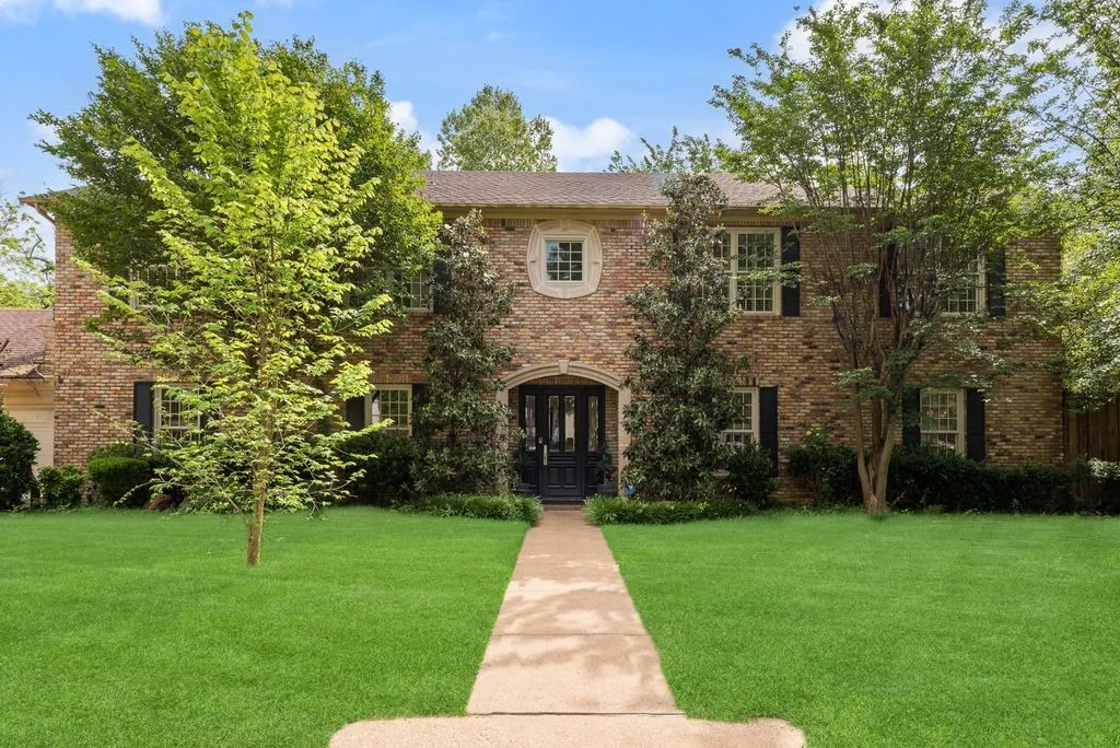 Two-story brick exterior featuring a stone arched entryway, symmetrical multi-pane windows with dark shutters, a decorative central oval window, and a paved walkway