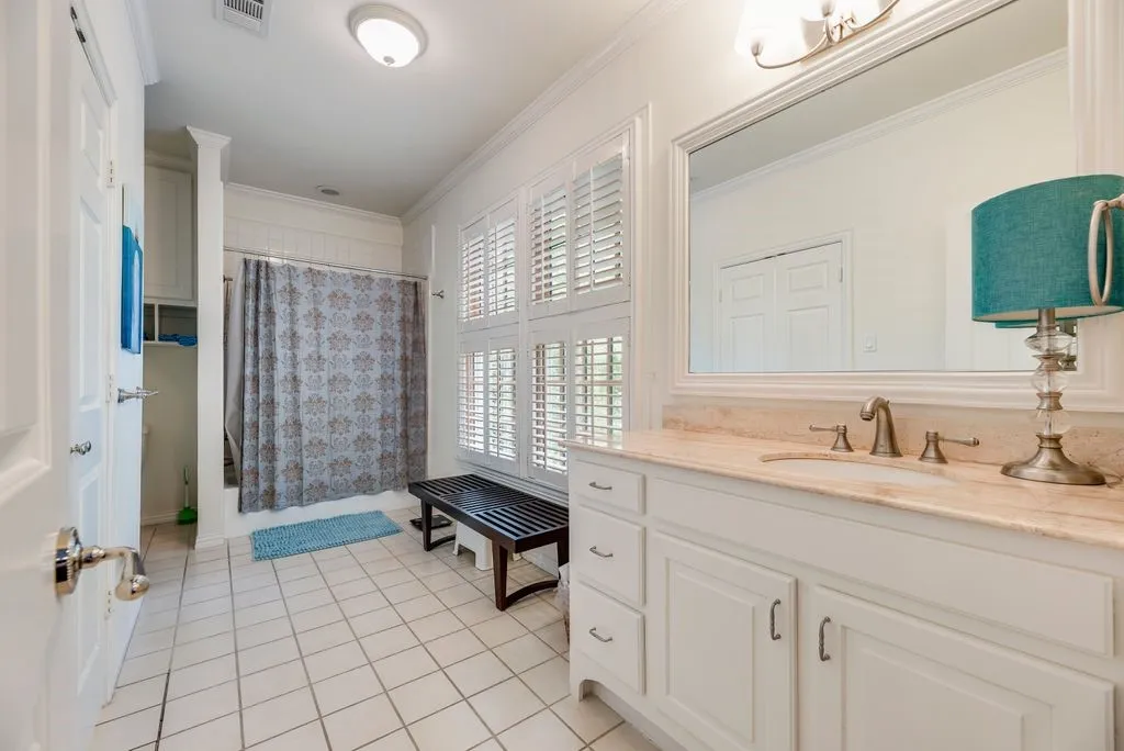 Bathroom featuring white cabinetry, light-toned stone countertop, large framed wall mirror, plantation shutters, and ceramic tile flooring