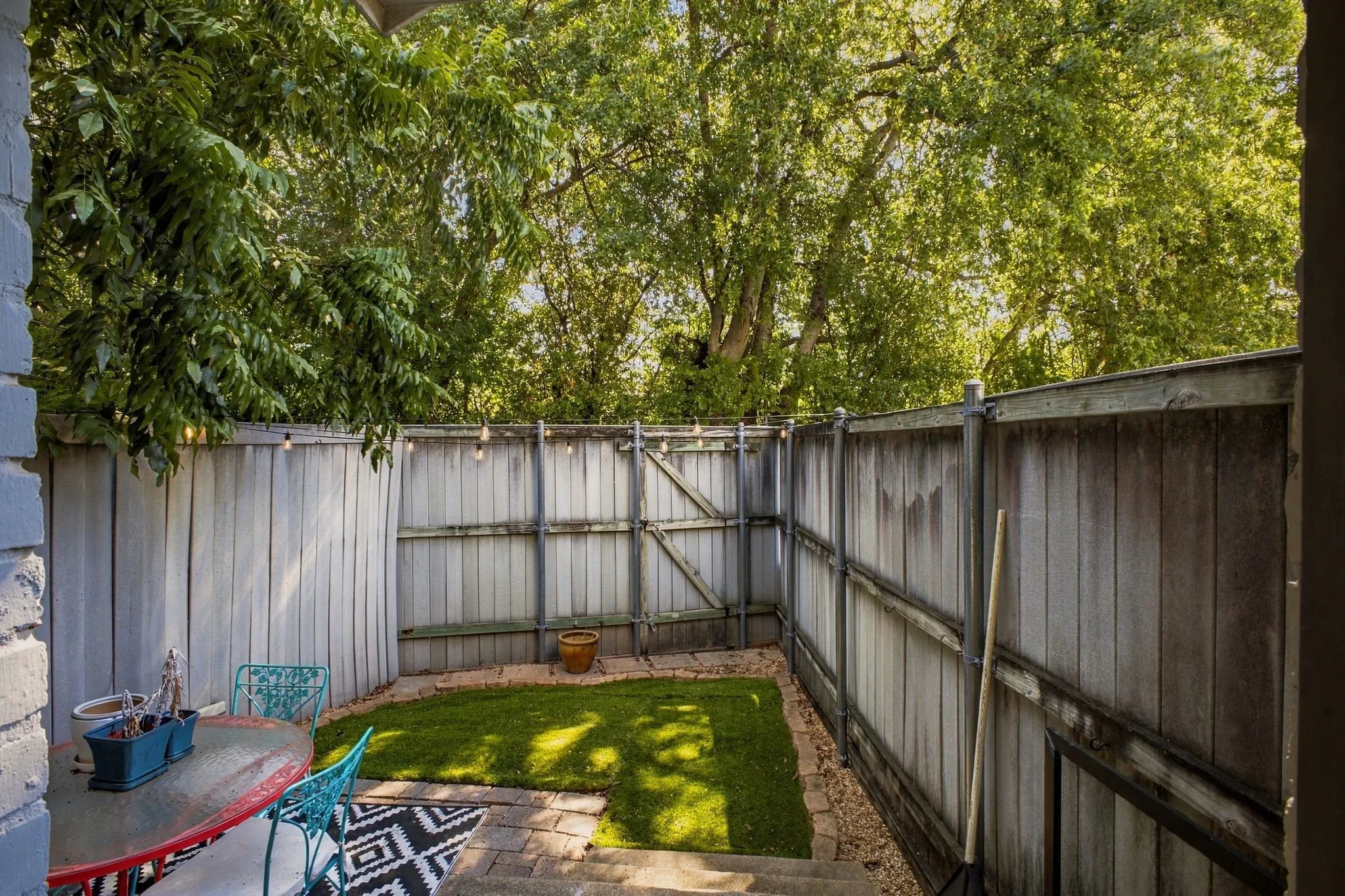 Enclosed outdoor space featuring a wood-panel fence, synthetic grass lawn, paver patio, and mature trees