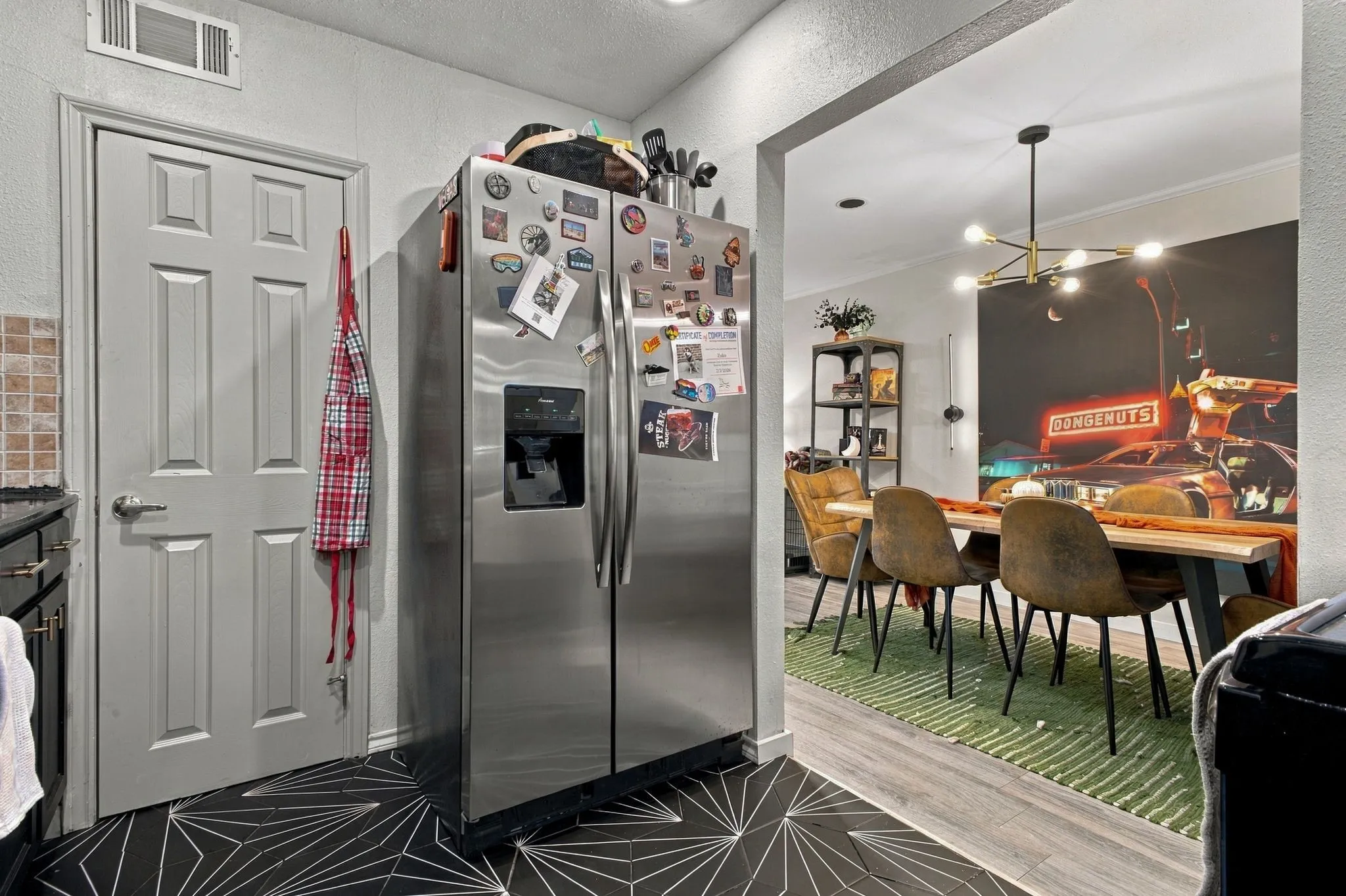 Stainless steel side-by-side refrigerator, geometric patterned floor tile, six-panel interior door with satin nickel hardware, and multi-arm chandelier lighting