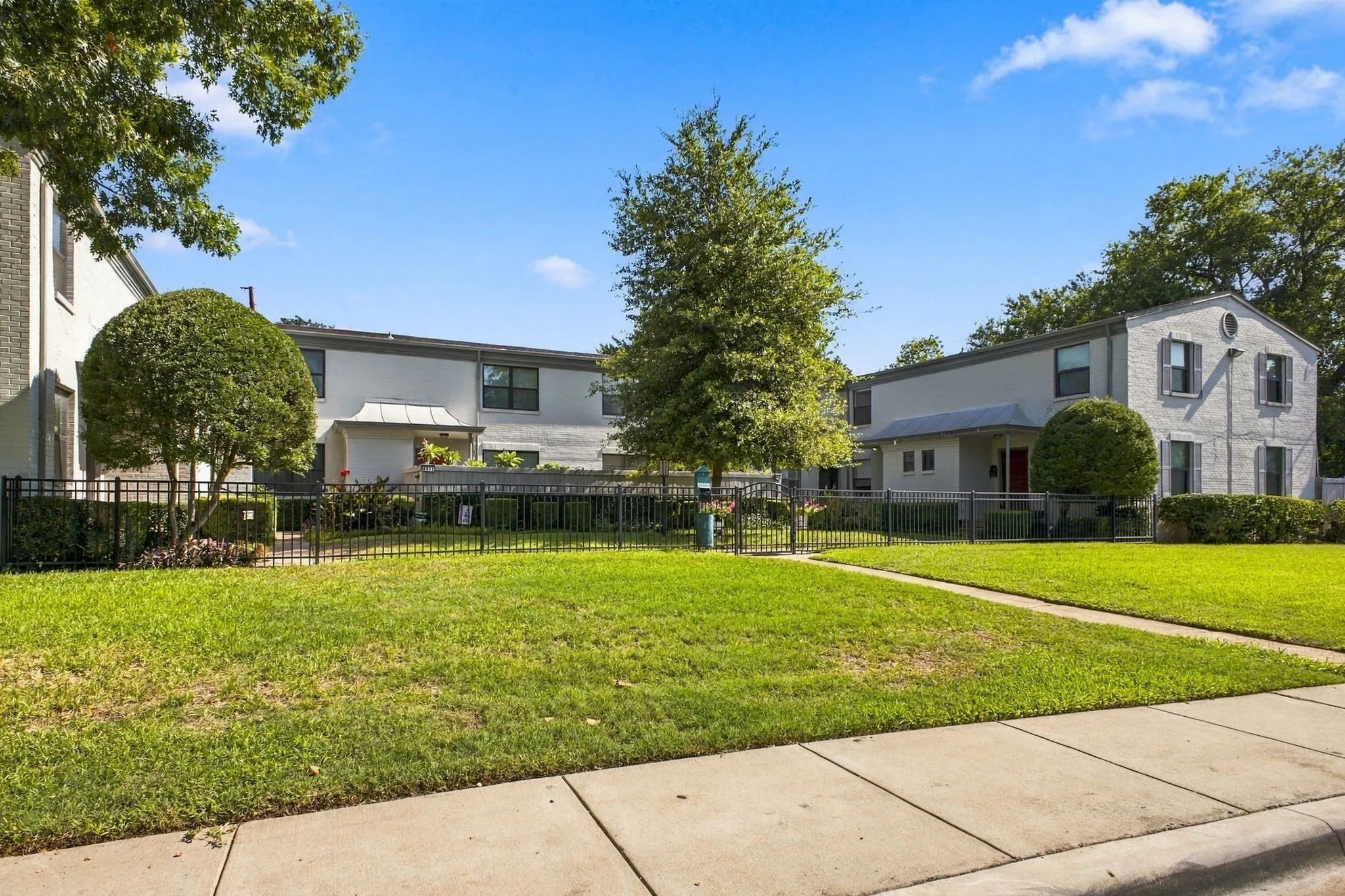 Multi-unit residential exterior featuring painted brick masonry, metal roof awnings, black metal perimeter fencing, and established landscaping