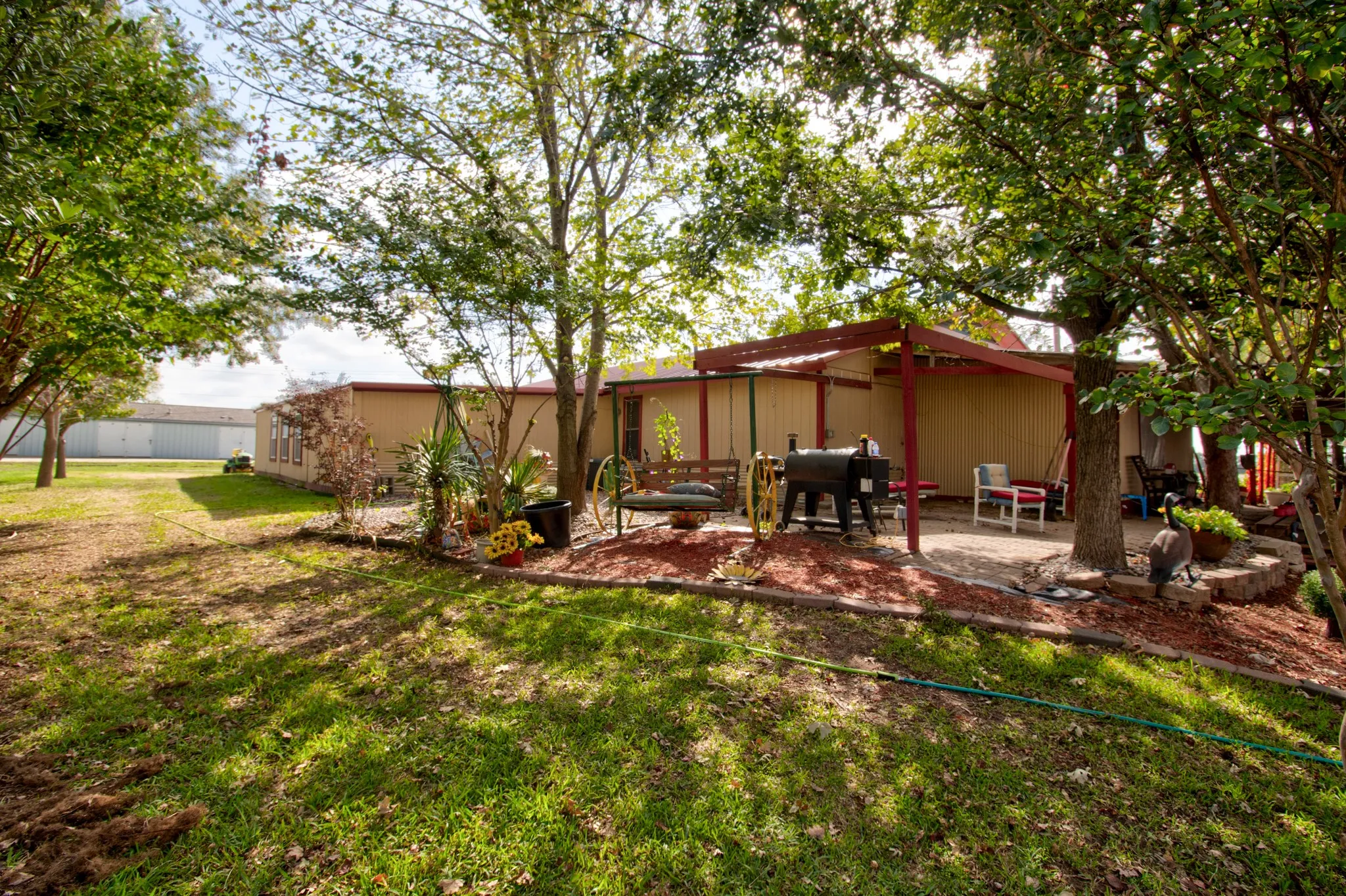 Rear exterior featuring a covered patio, paved hardscaping, mature trees, and landscaped garden beds