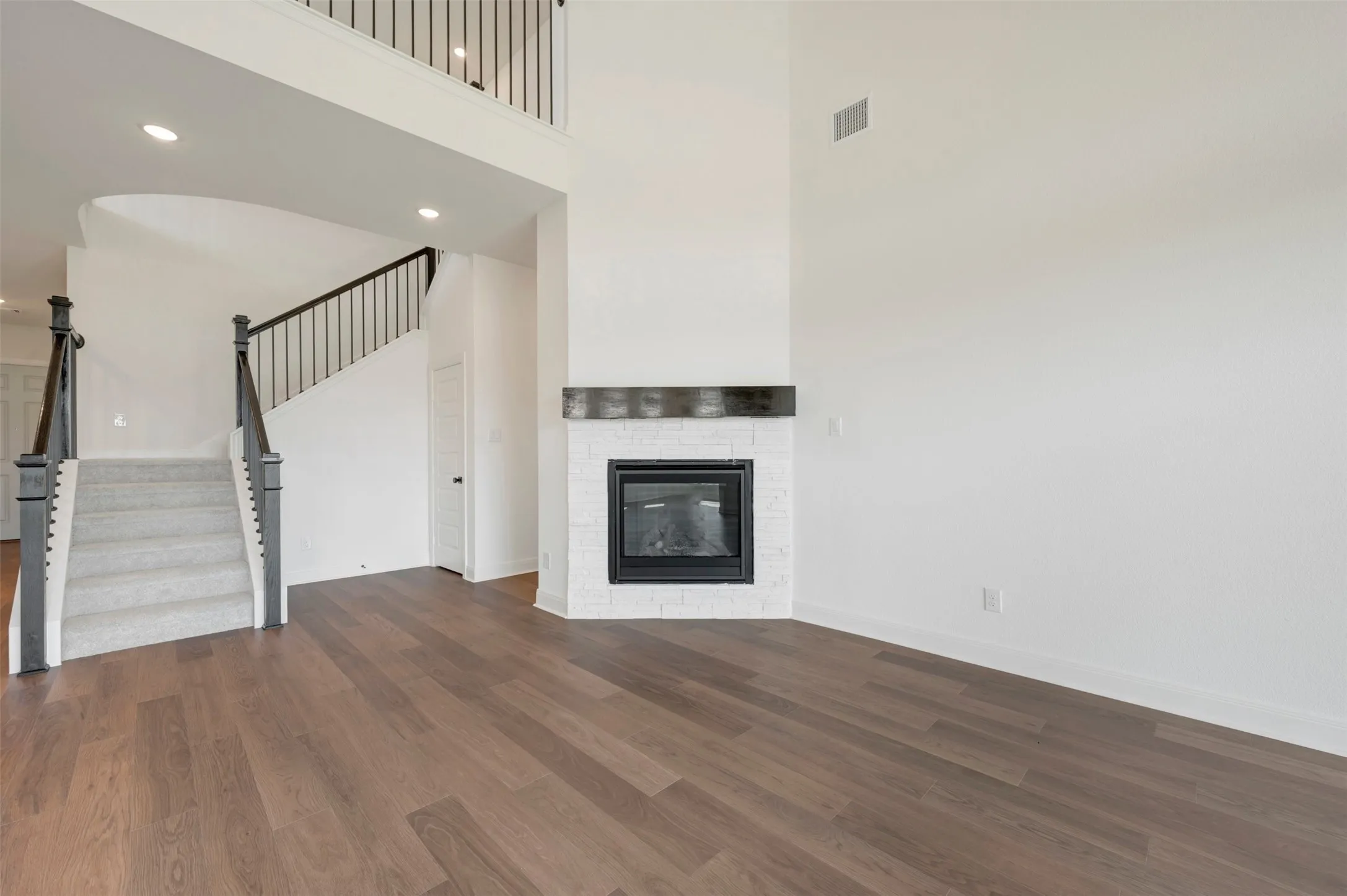 High-ceiling interior featuring wood-finish flooring, a white painted brick fireplace with a dark timber mantel, a staircase with metal balusters, and recessed lighting