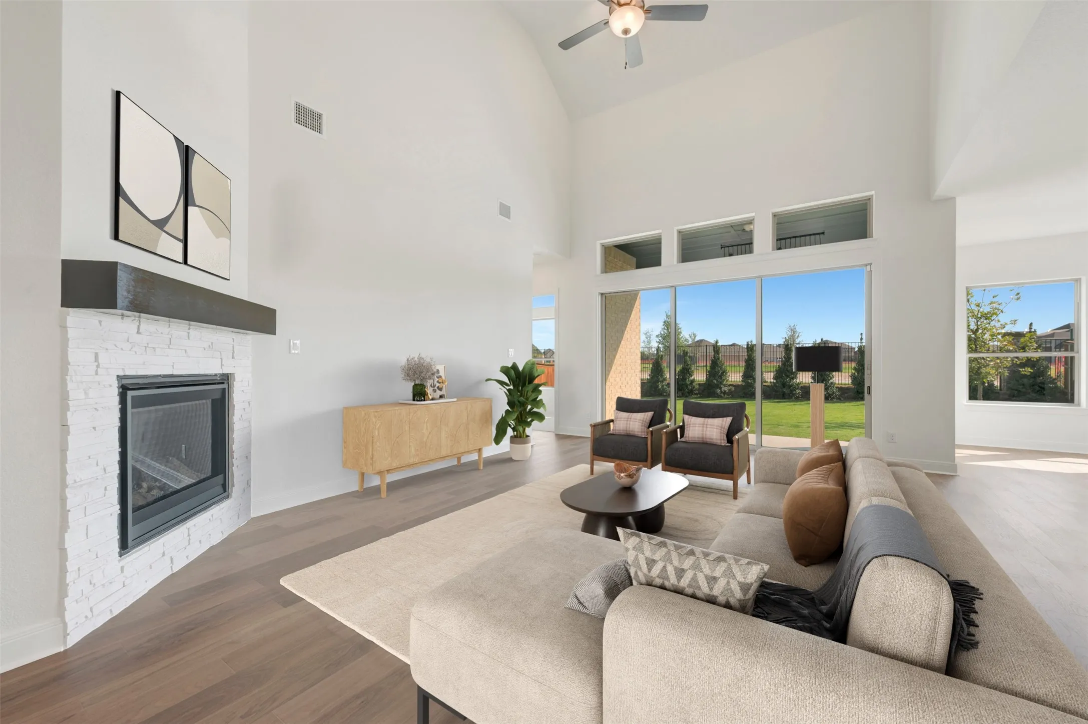 Vaulted ceiling with an integrated fan, floor-to-ceiling fireplace with painted brick finish and a prominent mantel, wood-finish flooring, and expansive windows featuring transom lights