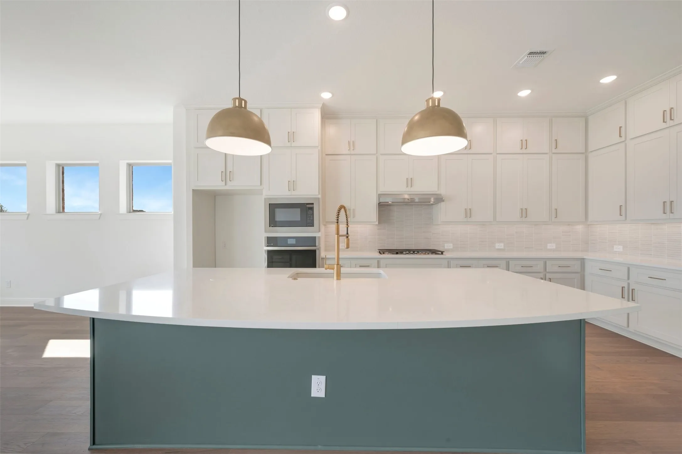 Kitchen island with stone-surface countertop, white cabinetry with silver-tone hardware, built-in microwave and wall oven, brass-finish faucet, and pendant lighting