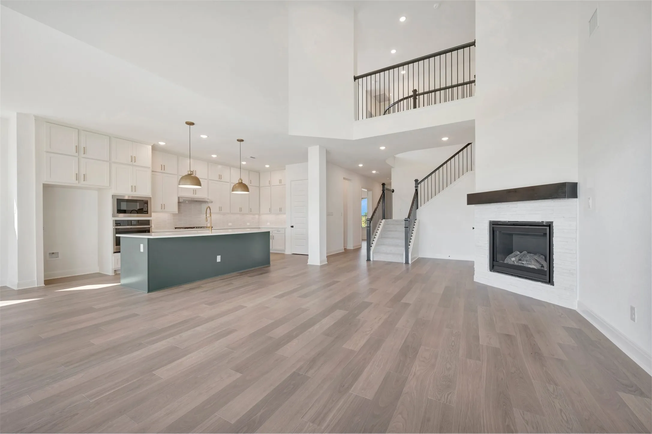 Open-concept interior featuring wood-finish flooring, a kitchen island with pendant lighting, built-in wall cabinetry, a fireplace with a masonry surround and dark mantel, and a staircase with decorative metal balusters