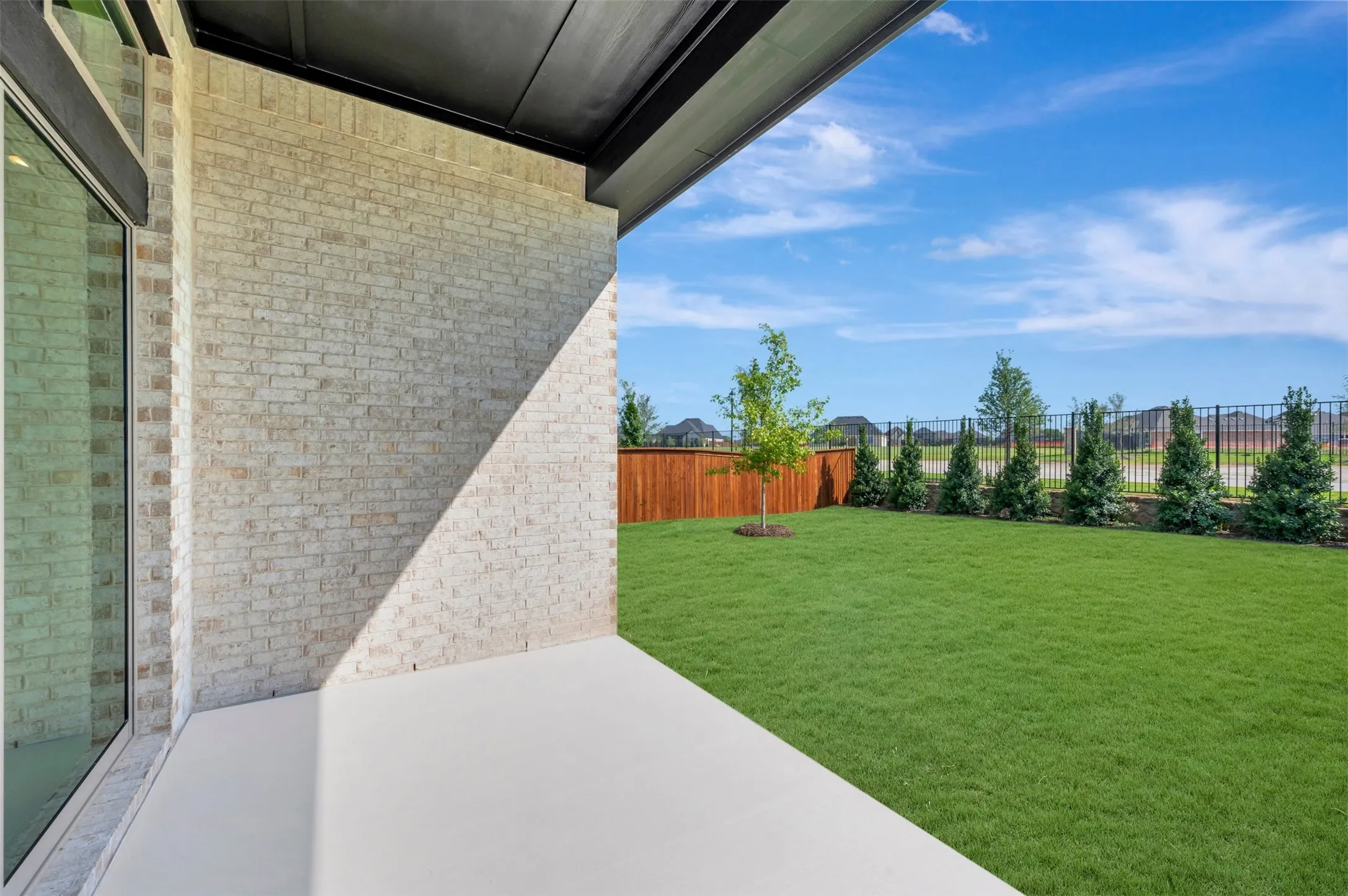 Covered patio featuring a light brick exterior wall, full-height glass door, manicured lawn, wood-panel fencing, and perimeter evergreen landscaping