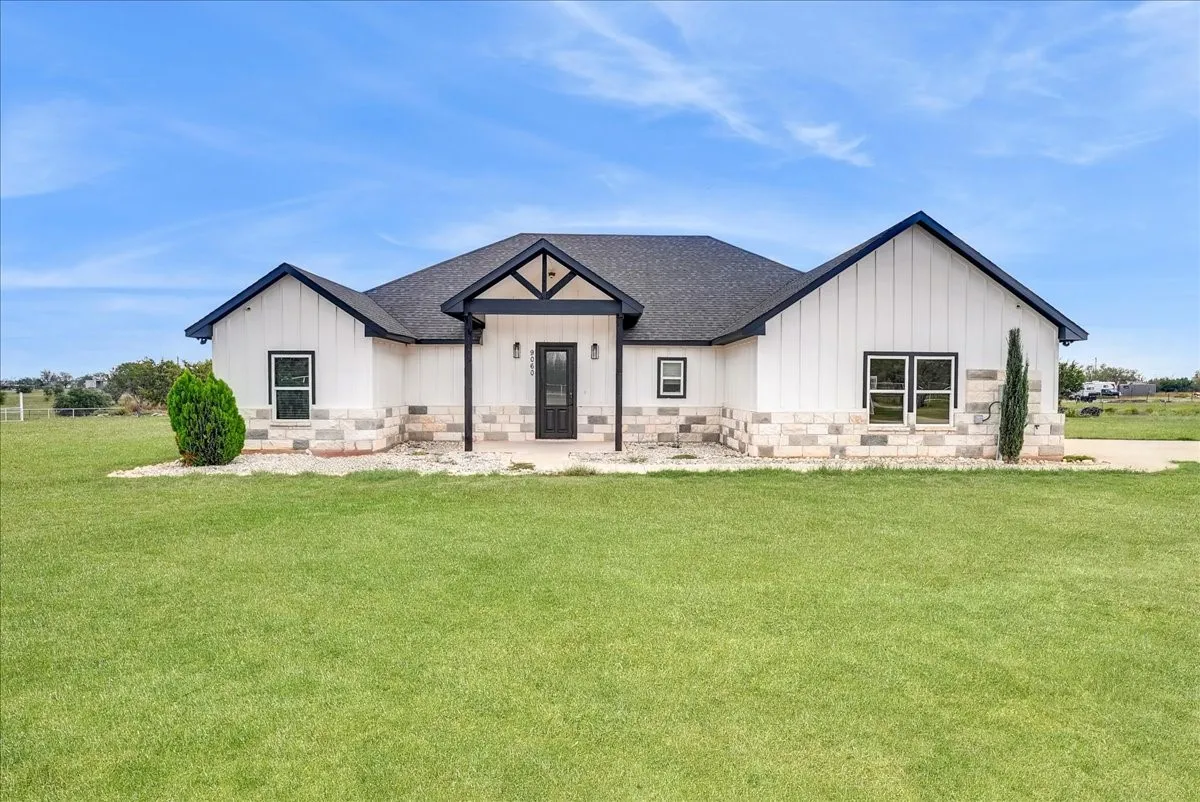 Single-story residence featuring white board-and-batten siding, natural stone veneer base, a covered front entryway with timber-frame detailing, and a shingled roof