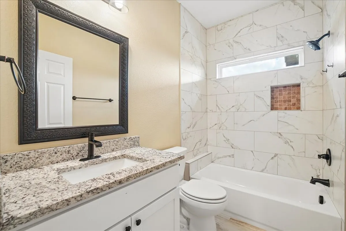 Bathroom featuring a vanity with stone-finish countertop, white cabinetry, rectangular under-mount sink, matte black fixtures, and a shower-tub combination with tiled surround and built-in niche