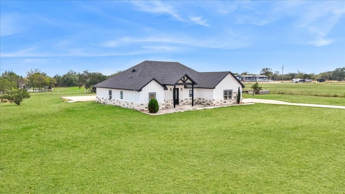 Single-story exterior featuring white vertical siding, stone masonry wainscoting, a dark shingle roof, and a covered front entry with timber-style accents, situated on expansive grounds