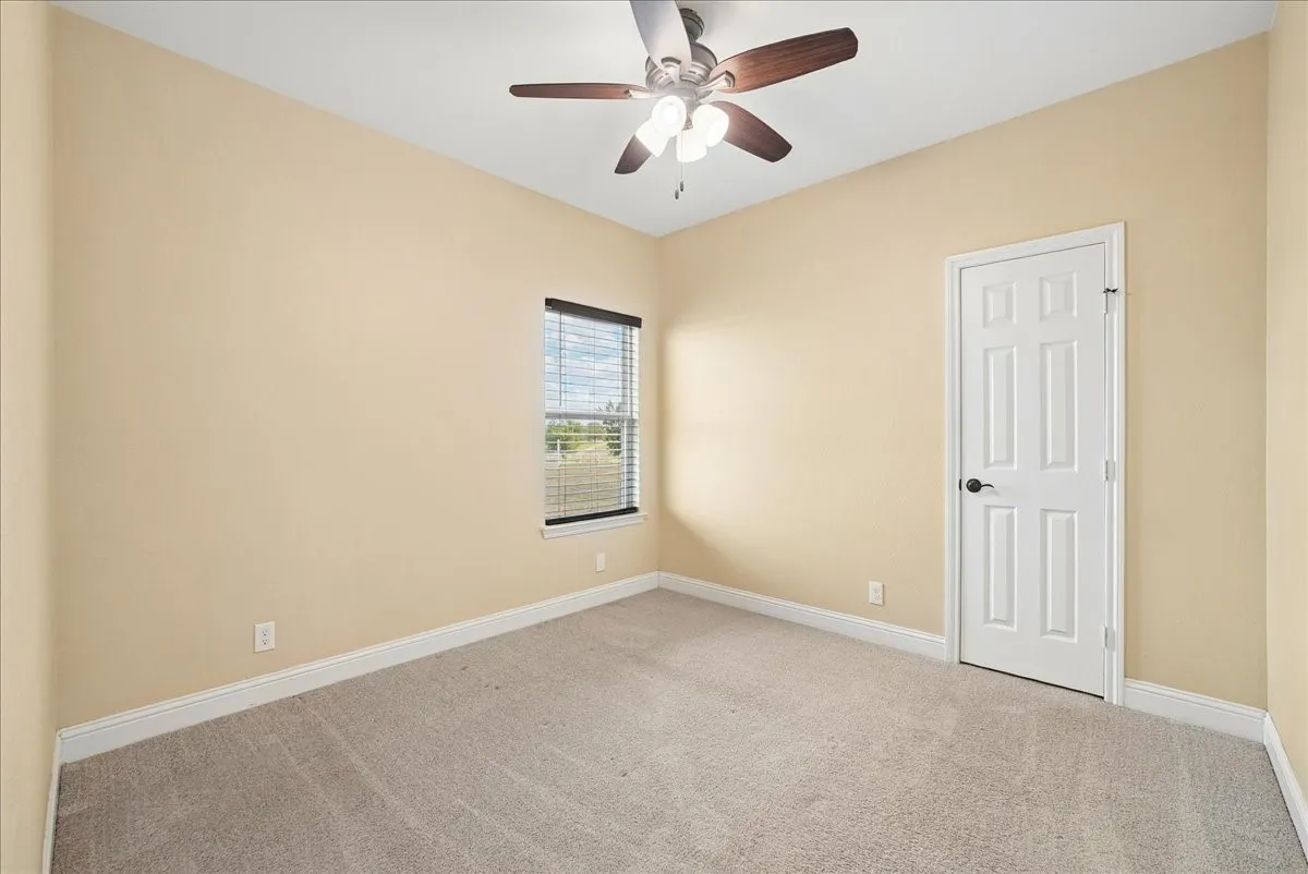 Neutral-toned interior space featuring neutral wall paint, wall-to-wall carpeting, a six-panel interior door, a single-pane window, and a multi-blade ceiling fan with an integrated light fixture