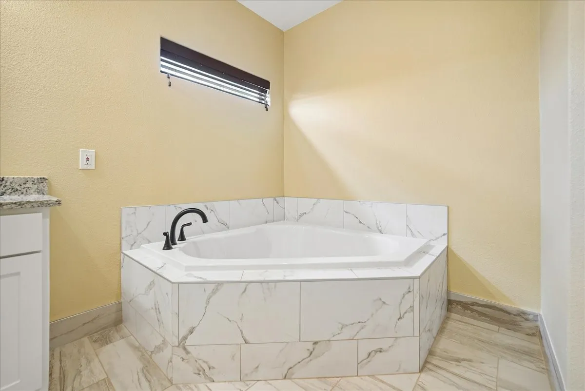 Corner soaking tub featuring a stone-look tile surround, white cabinetry, a stone countertop, and a matte black faucet