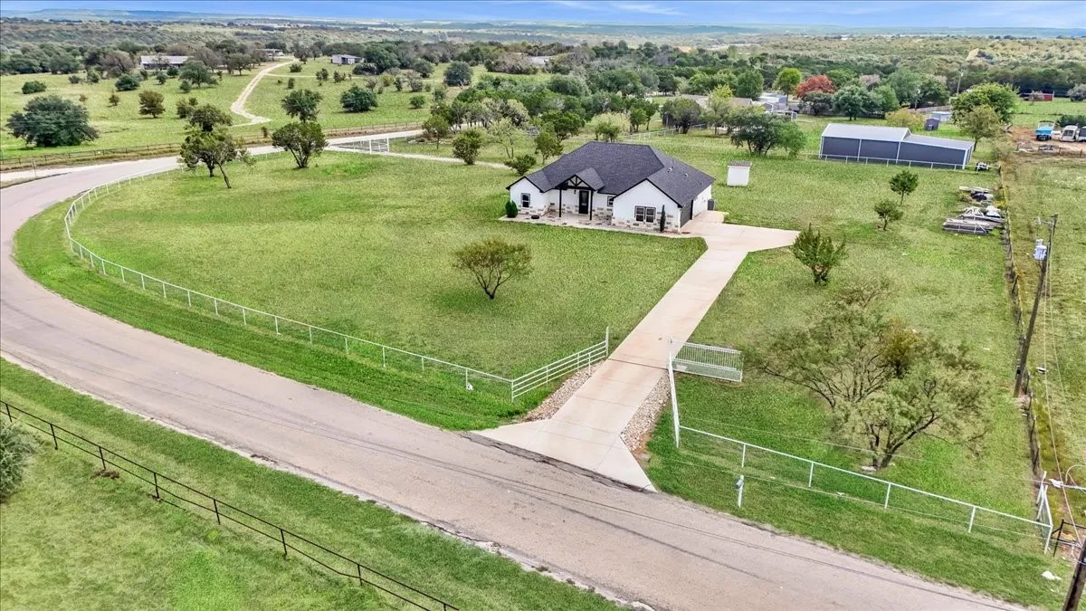 Single-story residence featuring a light-toned exterior, dark shingle roof, concrete driveway, perimeter metal fencing, and an auxiliary detached structure on a sprawling acreage lot