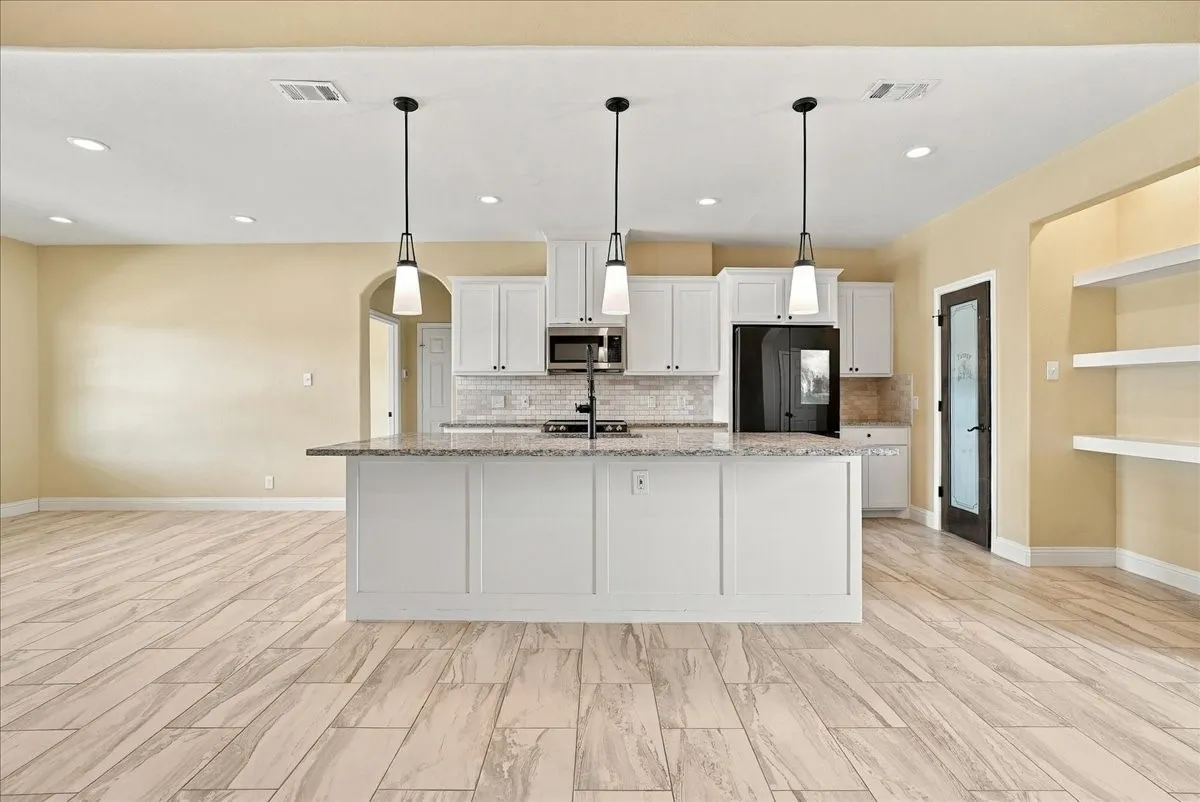 Kitchen featuring white shaker-style cabinetry, a stone-surfaced center island with an integrated sink, three overhead pendant lights, and light wood-finish tile flooring
