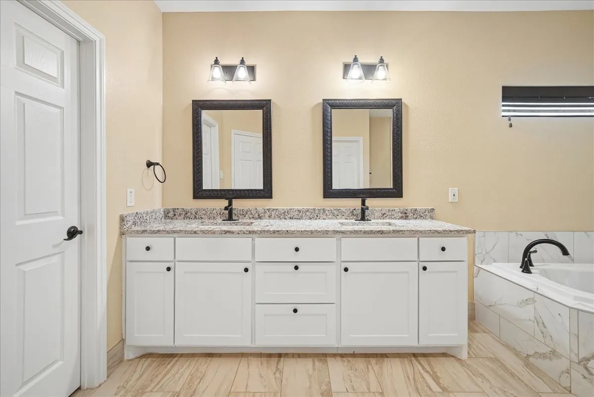 Double vanity with white shaker-style cabinetry, stone-finish countertop, dual under-mount sinks, dark-framed mirrors, and wood-finish flooring