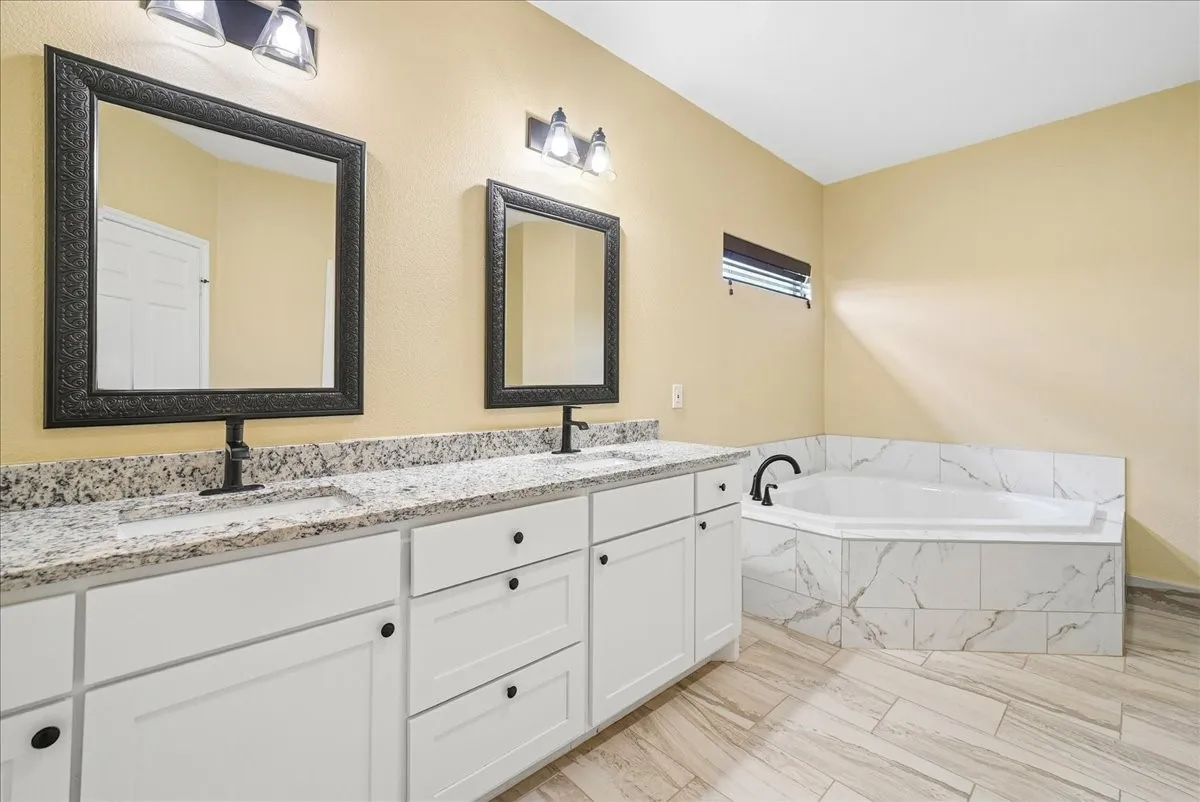 Dual vanity with light-toned stone-finish countertop, white shaker-style cabinetry, framed wall mirrors, corner soaking tub with surround tile, and tile flooring